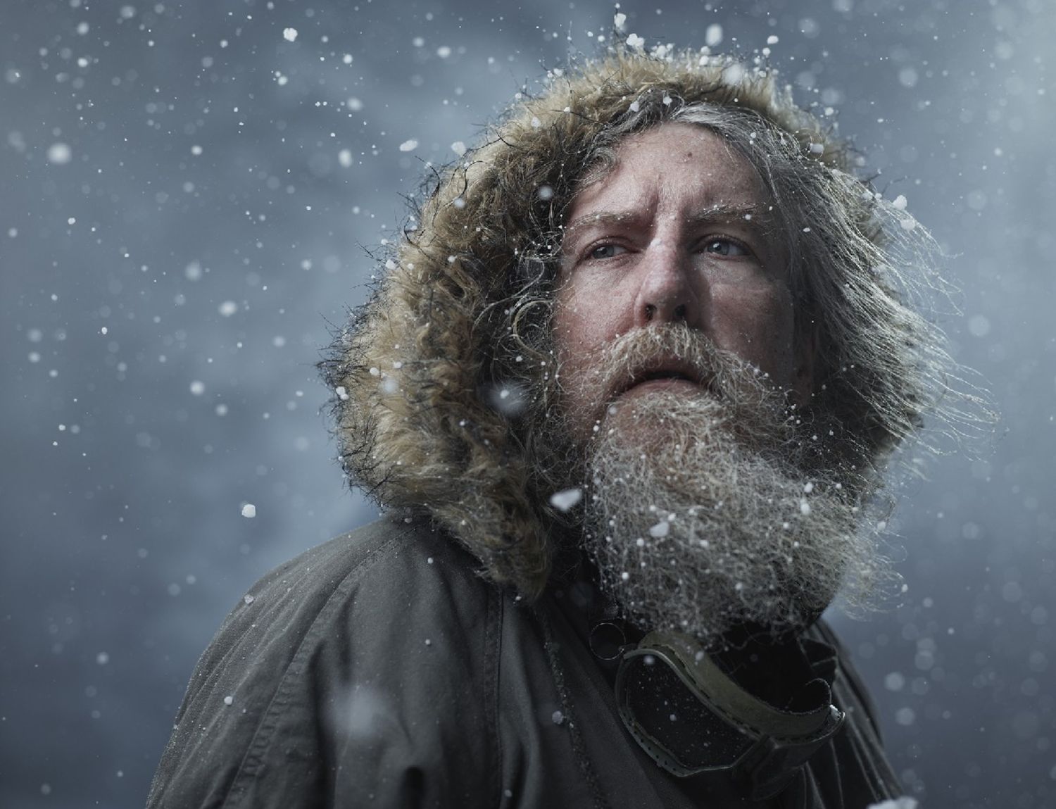 Dramatic portrait of a bearded man wearing a fur-lined parka looking up in a heavy snowstorm.