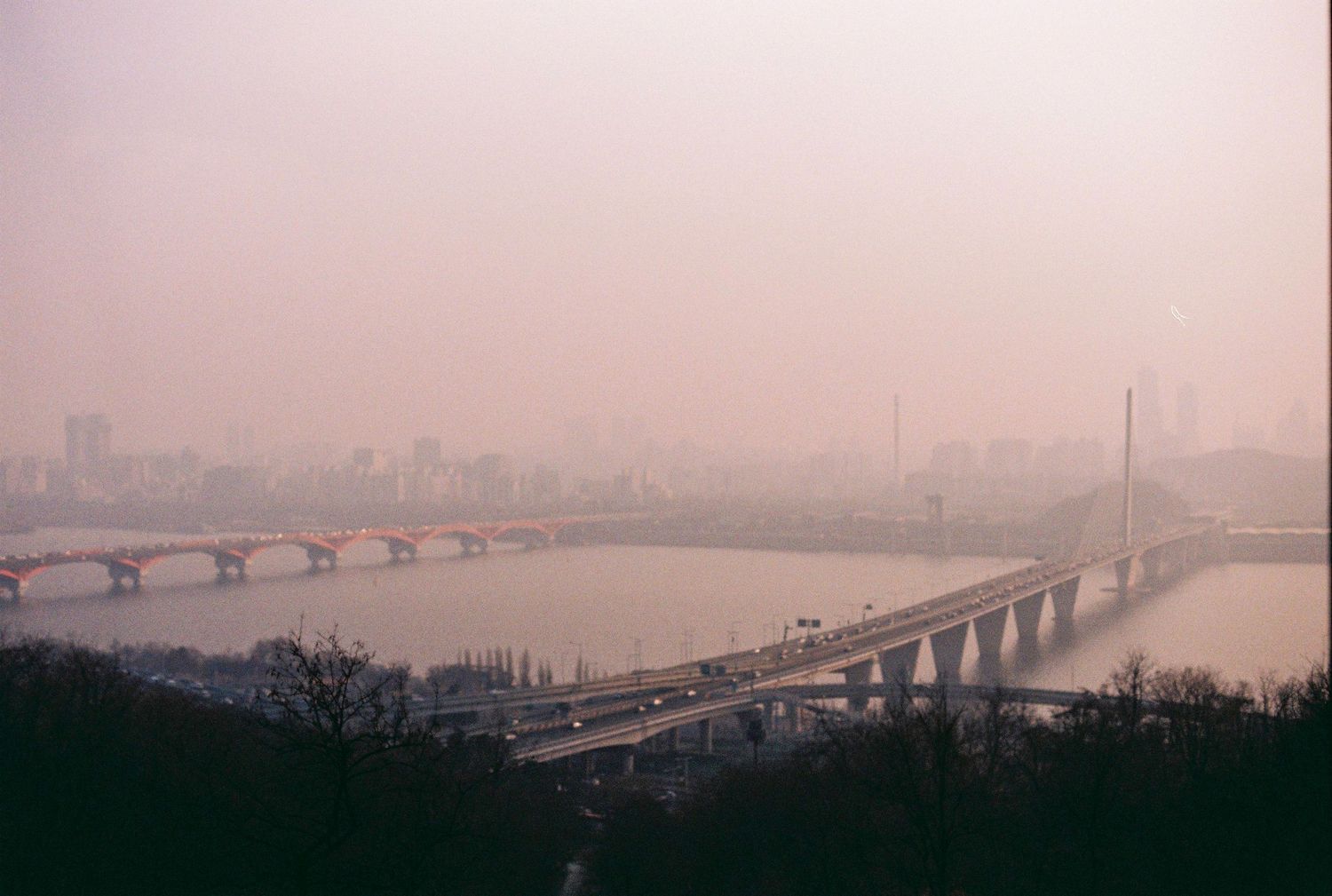 Hazy smog-covered cityscape of Seoul with a long bridge spanning a wide river at dusk in soft pink tones.