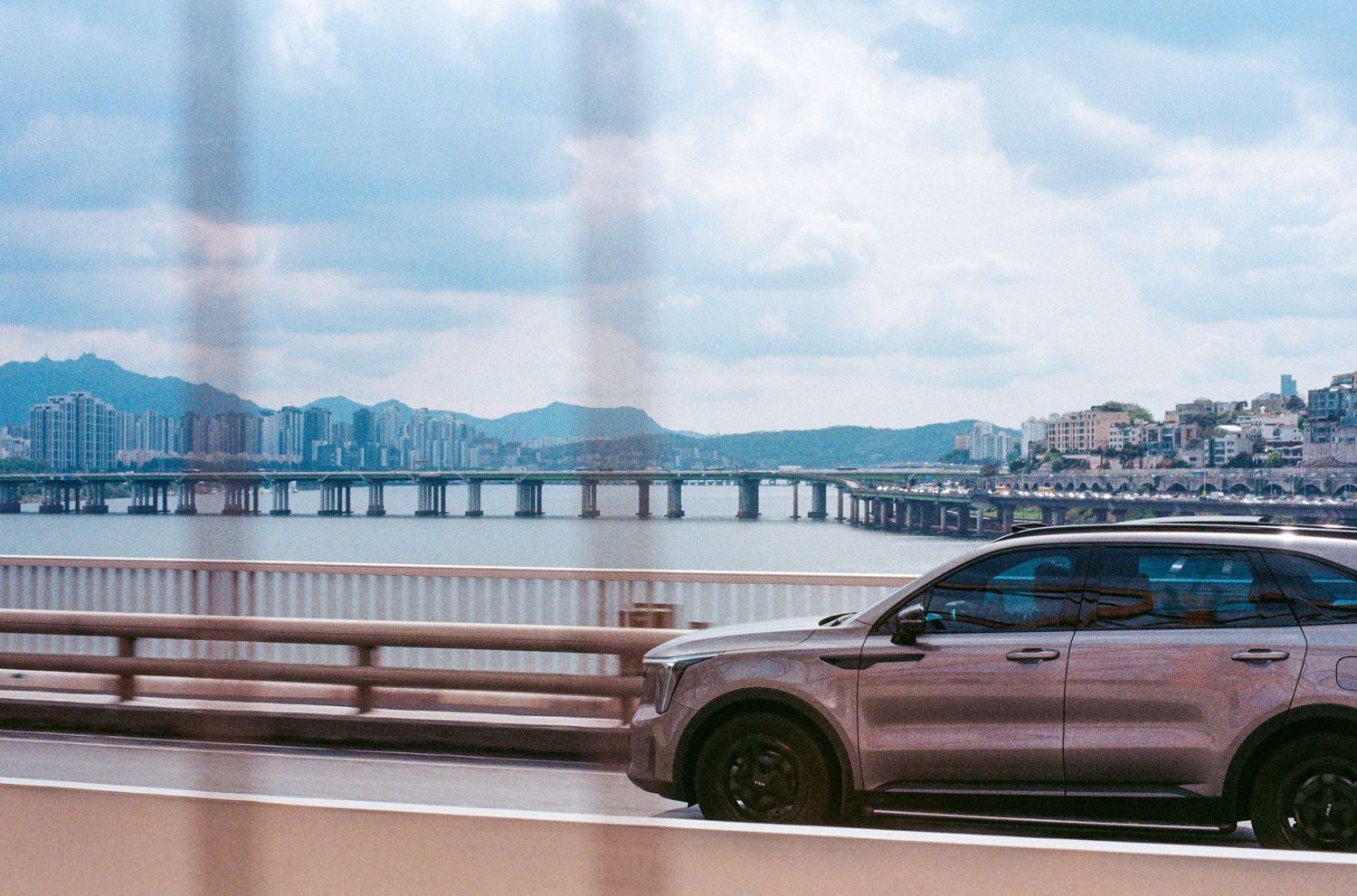 Silver SUV driving on bridge with city skyline and river in background under cloudy sky.