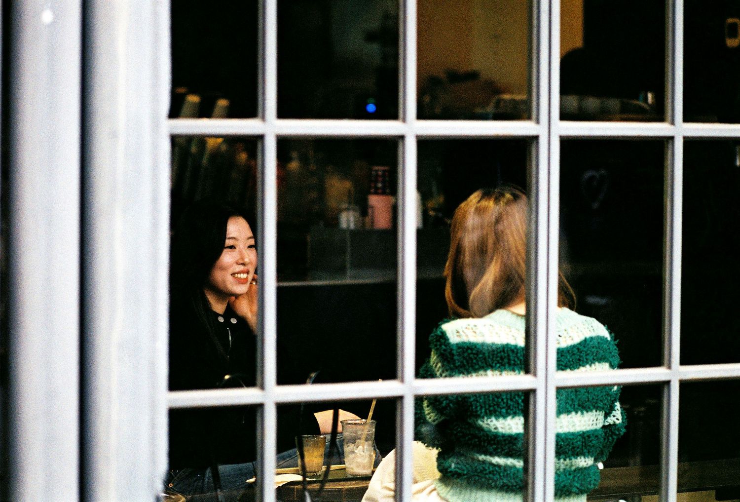 Two women conversing at a cafe table, viewed through a white-framed window in Seoul.