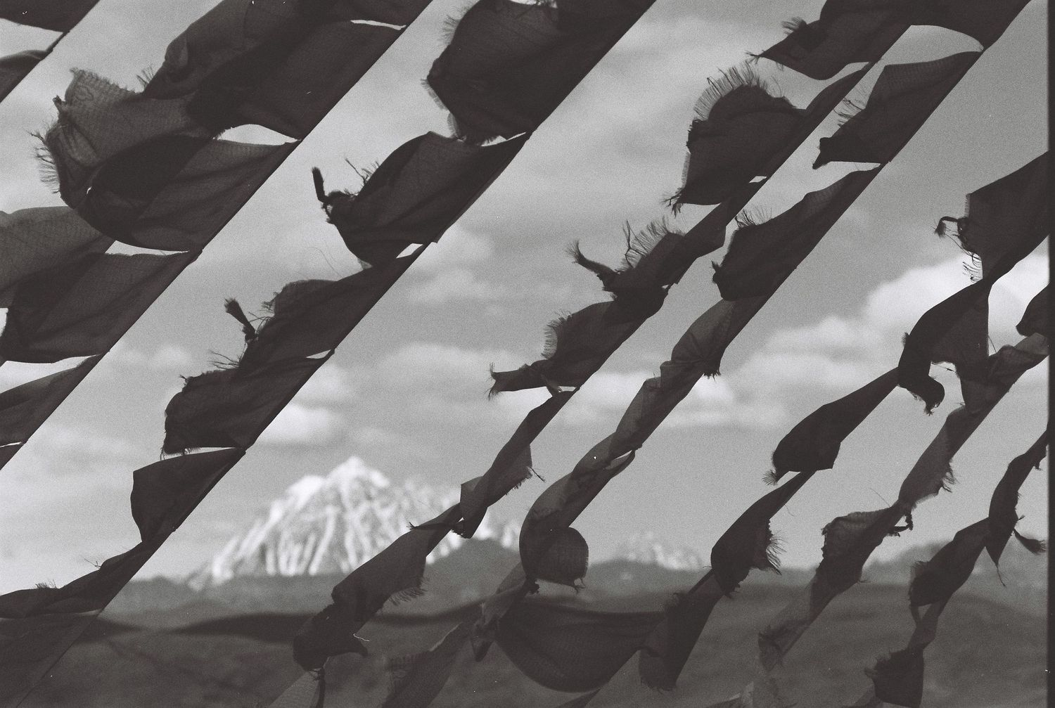 Black and white photo of tattered prayer flags blowing in wind with a mountain peak behind. It is a tibet mountain in China.
