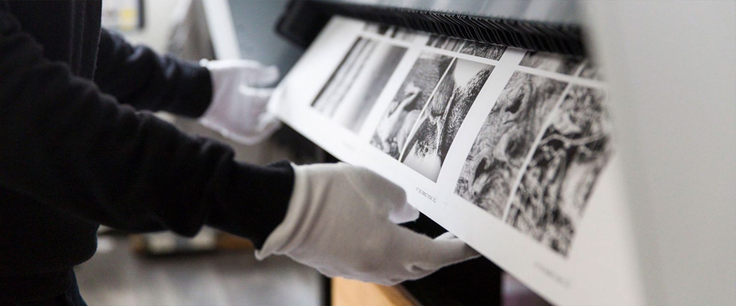 Gloved hands carefully handling large black and white photographic prints on a wide-format printer in a studio.