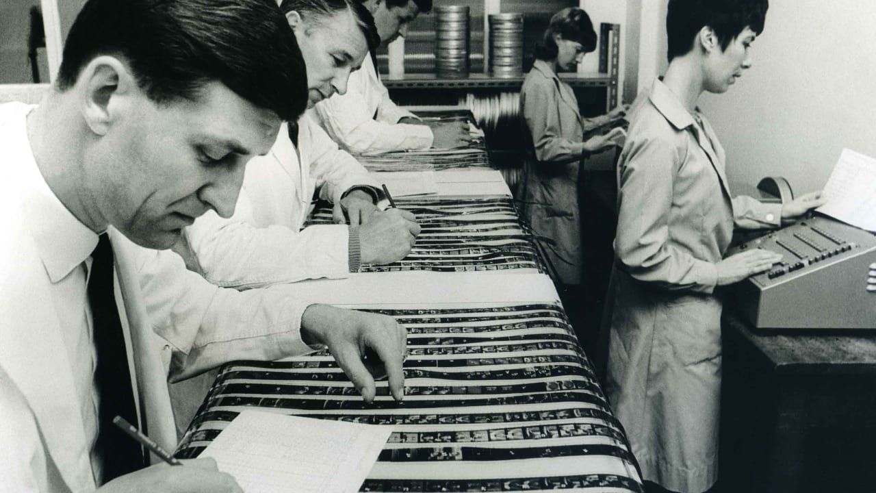 Black and white photo of workers examining film strips on a light table in a laboratory.