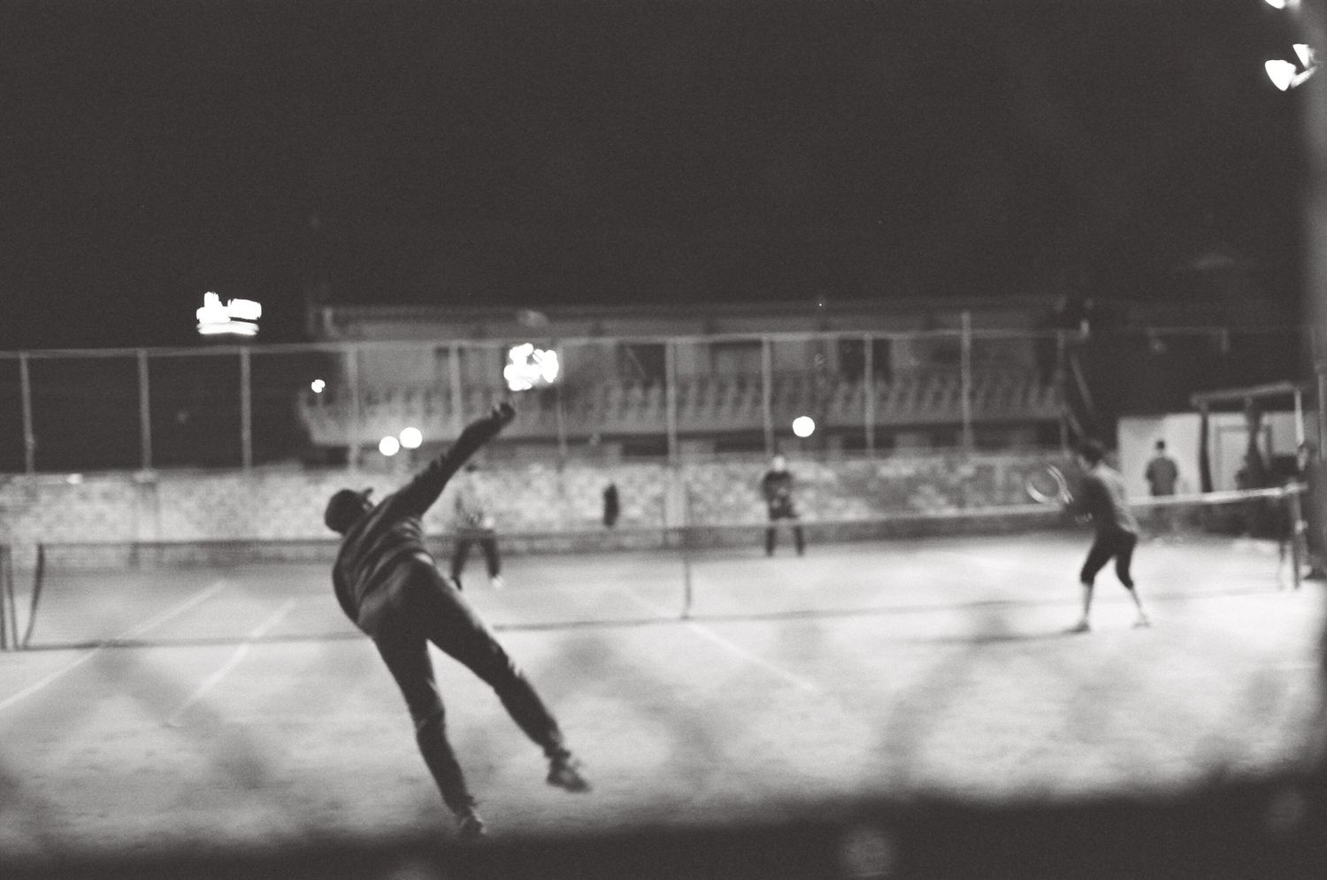 Black and white photo of a tennis player serving under night lights on an outdoor court.