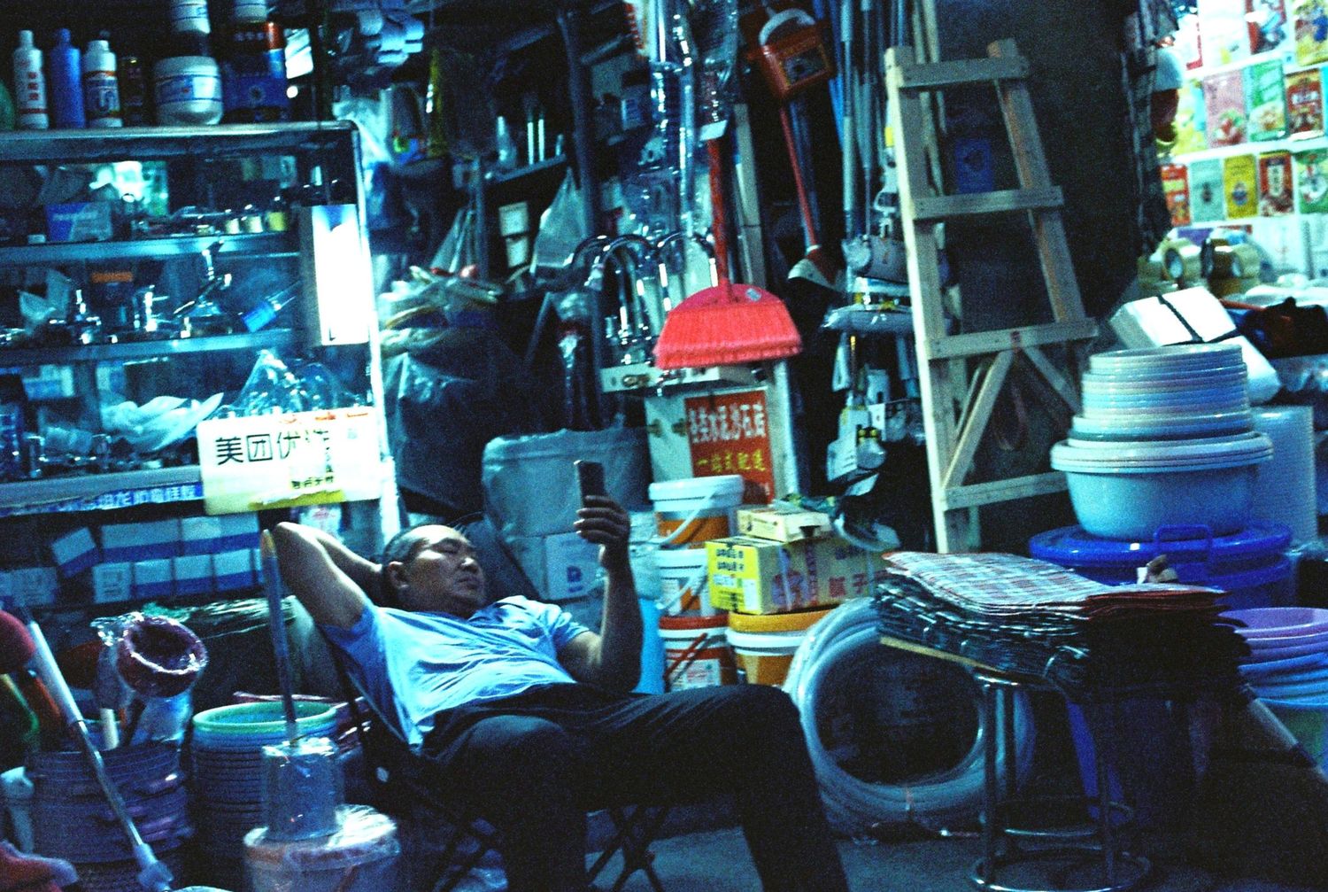 Man relaxing in China among densely packed colorful merchandise in a small cluttered shop.