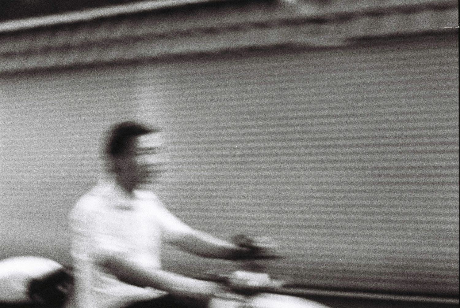 Blurry black and white photo of a man riding a motorcycle past a corrugated metal shutter wall.