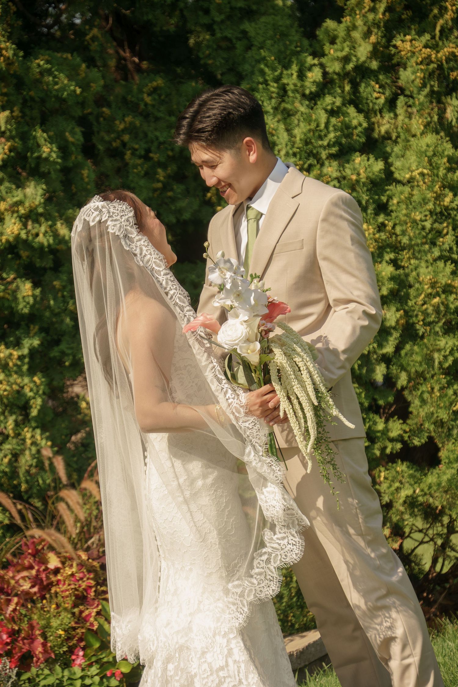 Bride and groom share a romantic moment outdoors, she in lace veil and he in beige suit holding a white bouquet.