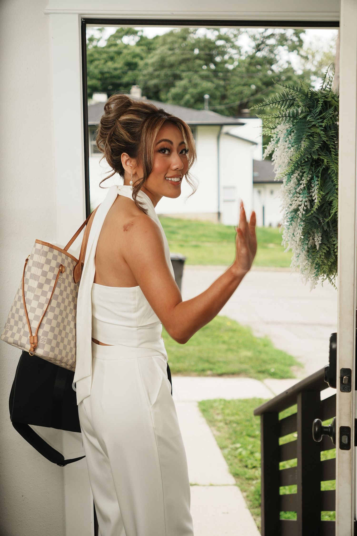 Woman in white halter outfit with Louis Vuitton bag waving goodbye at front door with green wreath.