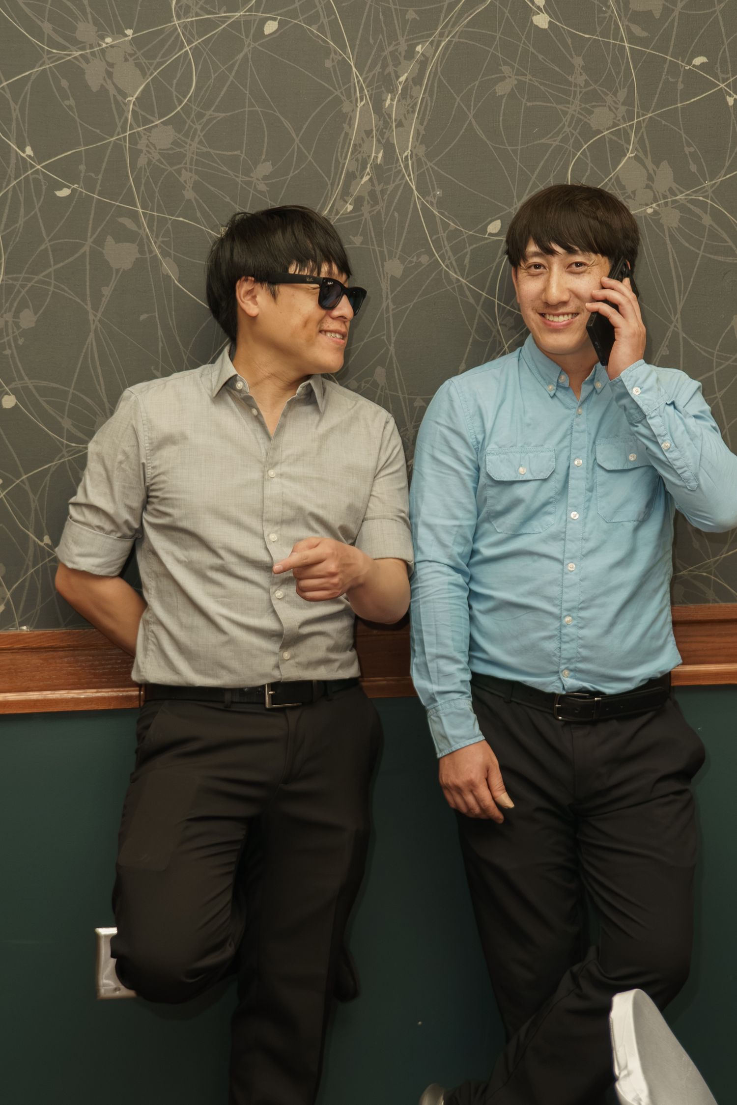 Two young Asian men lean against chalkboard wall, one wearing sunglasses, other talking on smartphone.