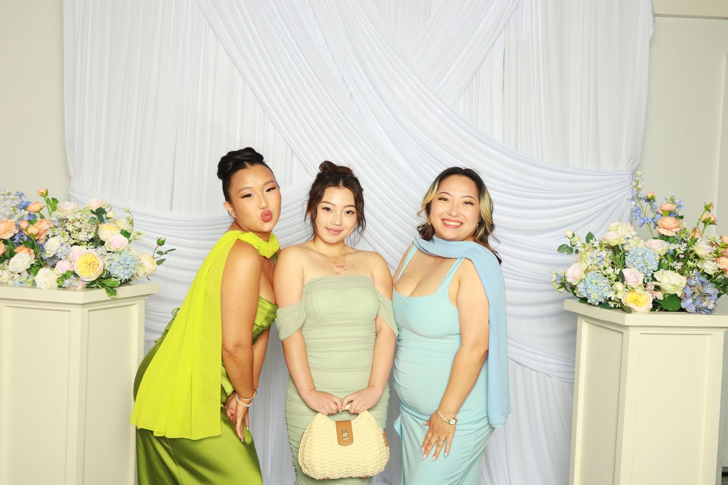 Three women in green and blue dresses pose at floral wedding backdrop.