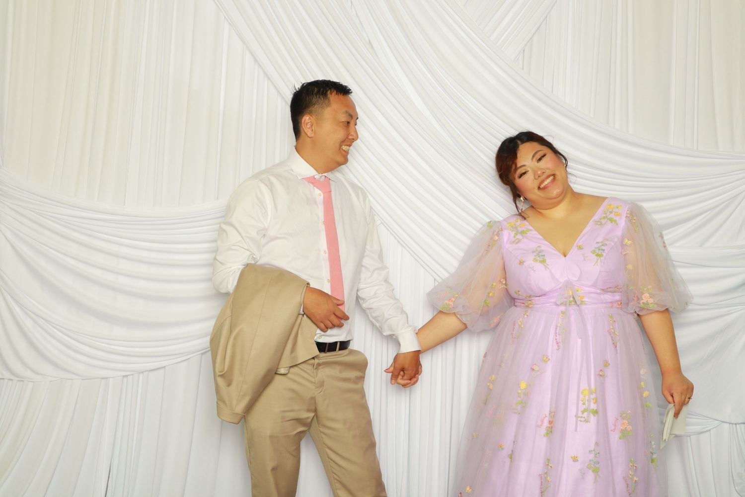 Couple holding hands in front of white draped backdrop at wedding celebration.