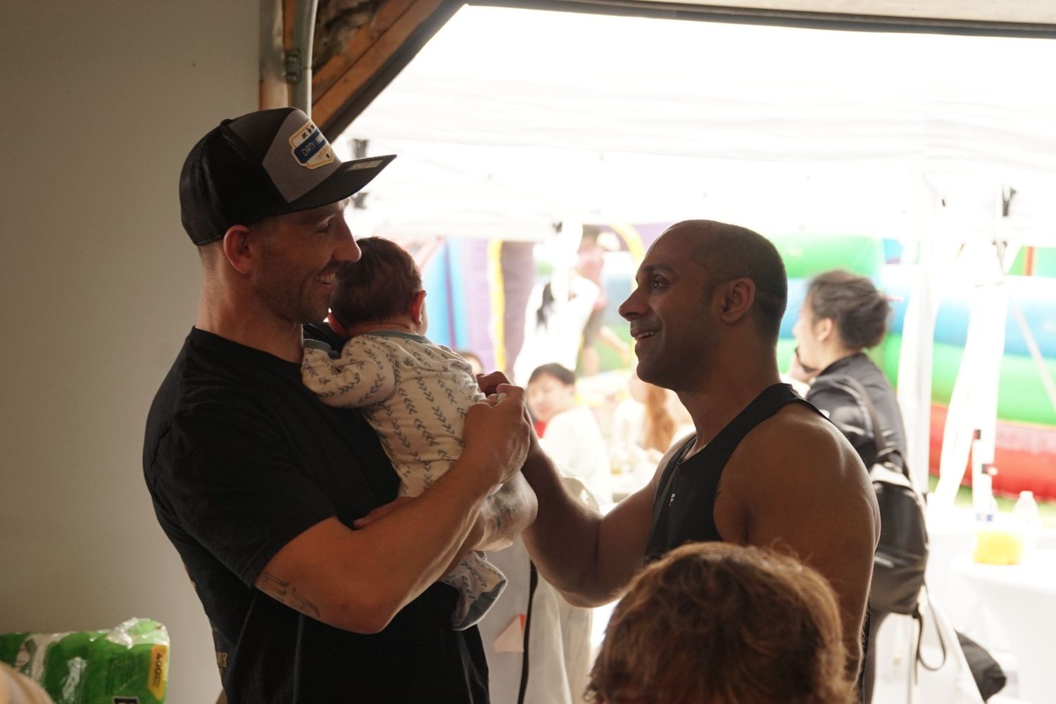 Two men conversing indoors, one holding a baby, in a casual social gathering setting.