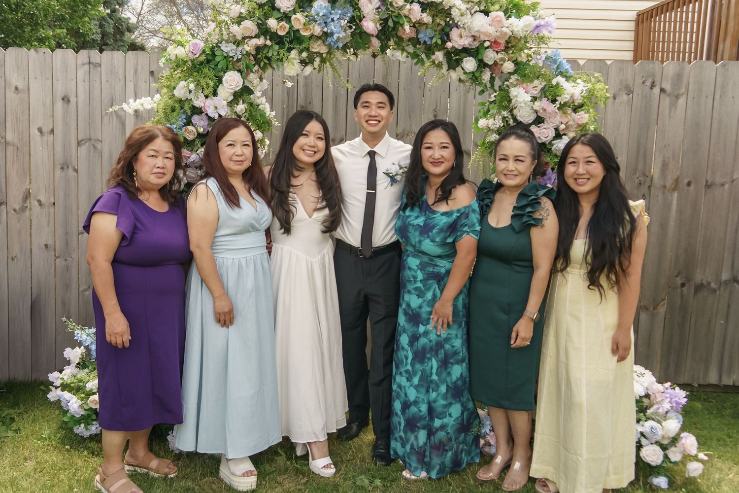 Bride and groom with six female family members posing under floral wedding arch outdoors.