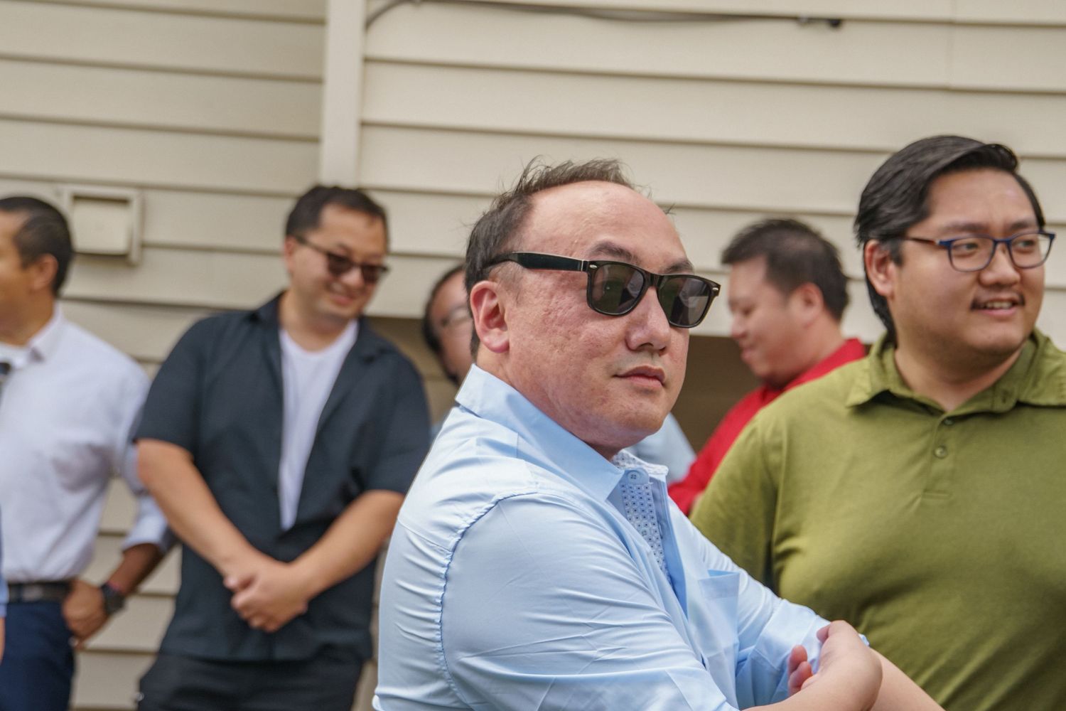 Man in light blue shirt with sunglasses stands with arms crossed at outdoor gathering.