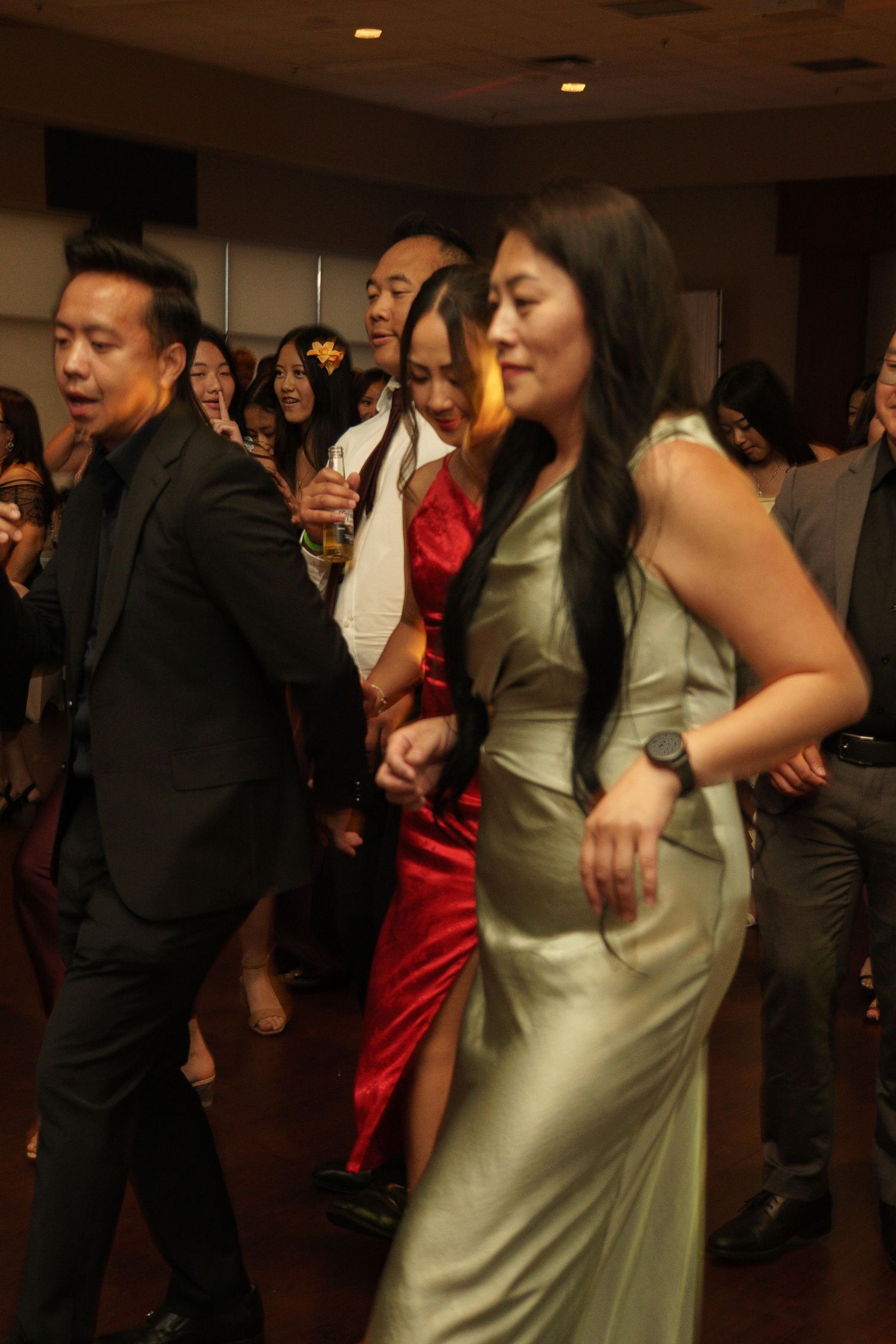 Guests in elegant formal attire, including green satin and red dresses, dance together at an indoor event or wedding reception.
