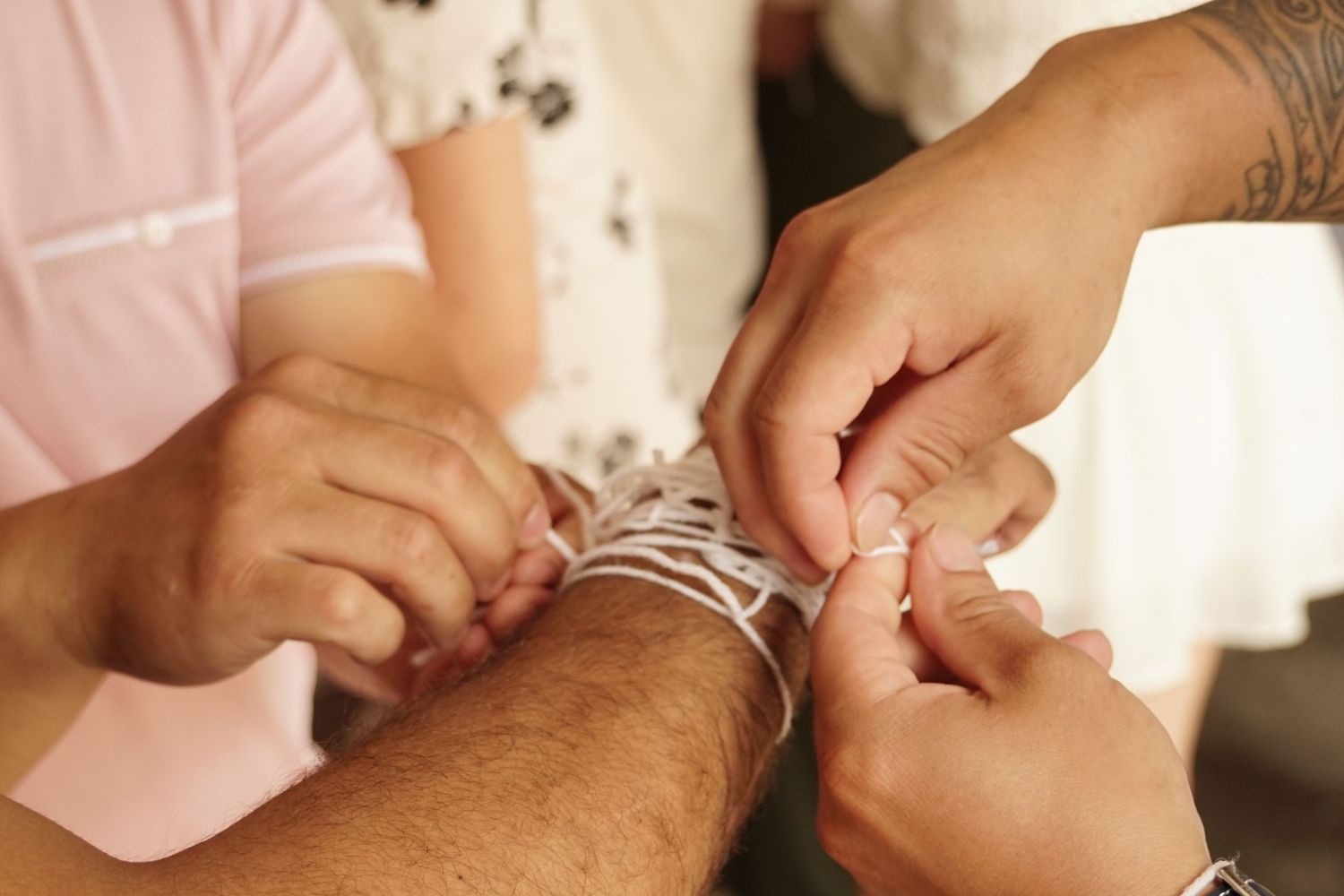 Close-up of multiple hands fastening a delicate white bracelet on a wrist, with a tattooed arm visible.