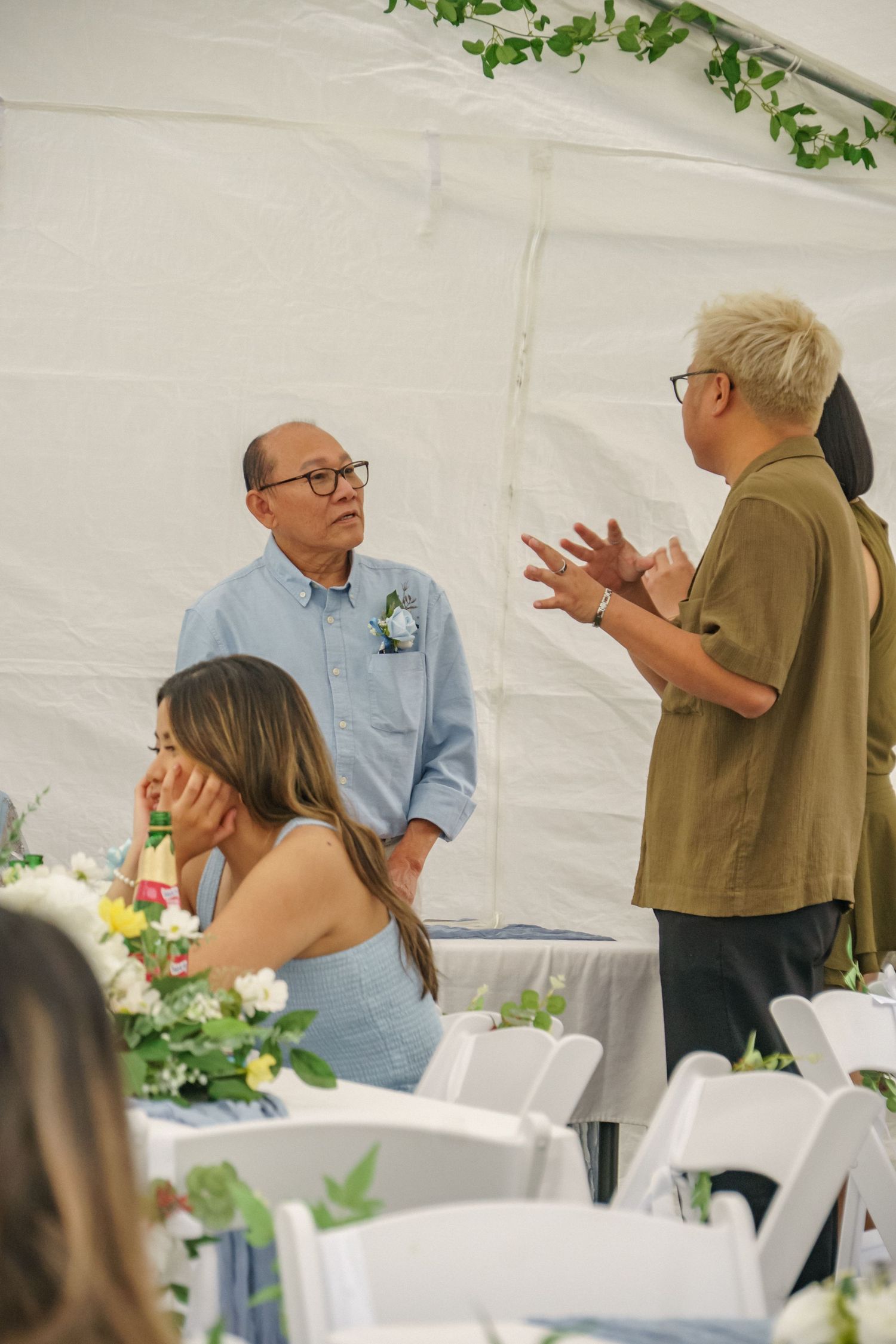 Two men in glasses converse at an elegant outdoor wedding reception under a white tent with floral centerpieces.