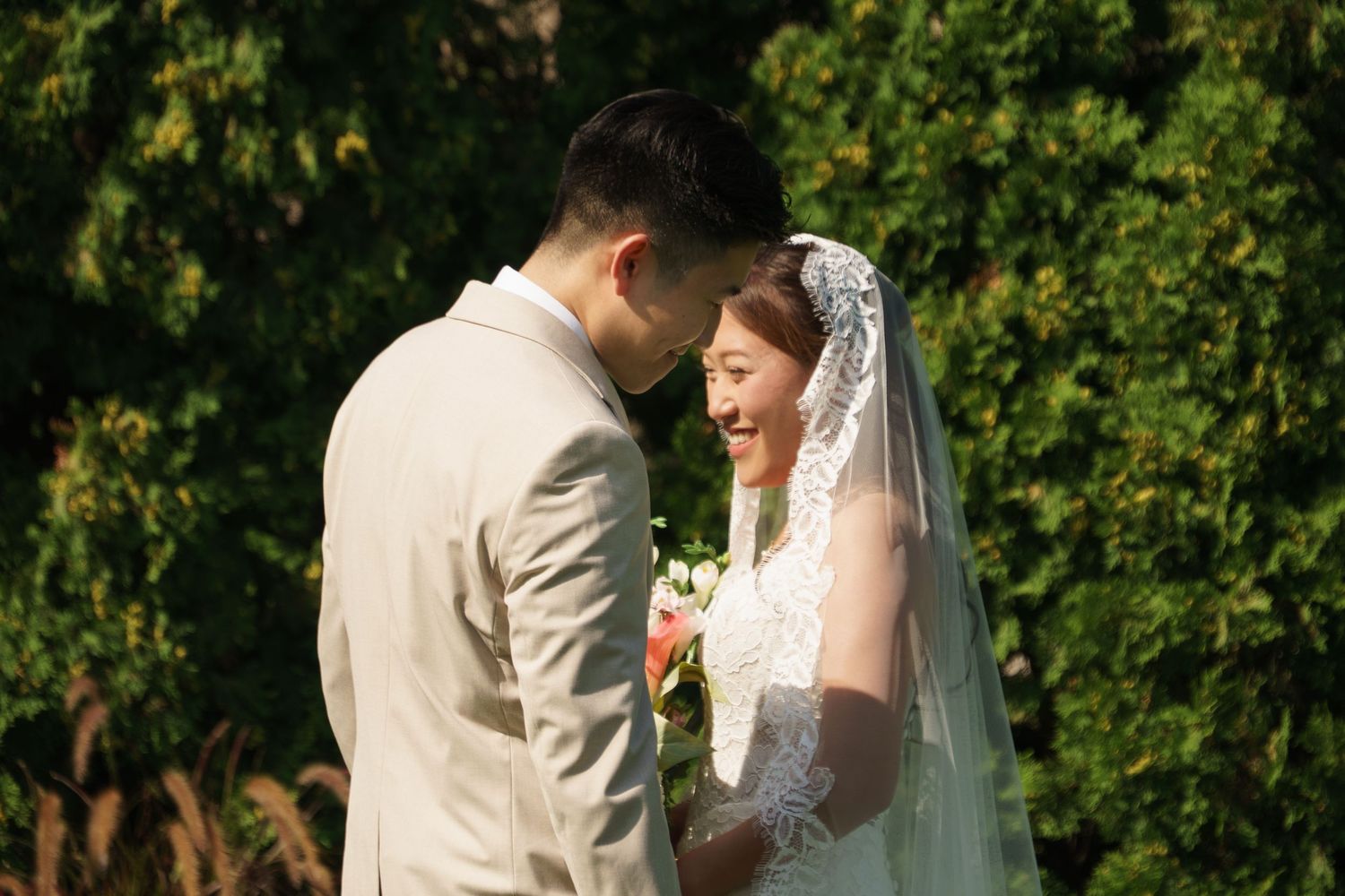 Asian bride and groom share a tender moment outdoors, bride smiling in lace veil holding bouquet.
