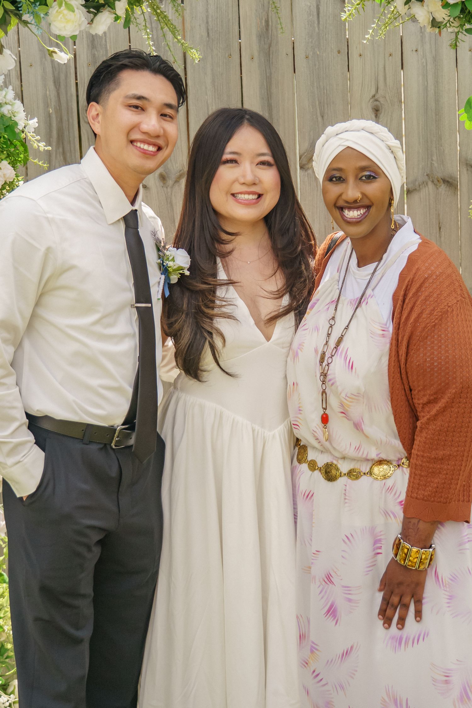 Couple in wedding attire smiles with guest in hijab at outdoor garden celebration.