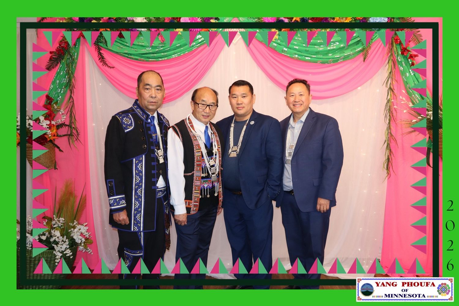 Four men in formal attire pose together in front of a decorative pink and green backdrop at a celebratory event.