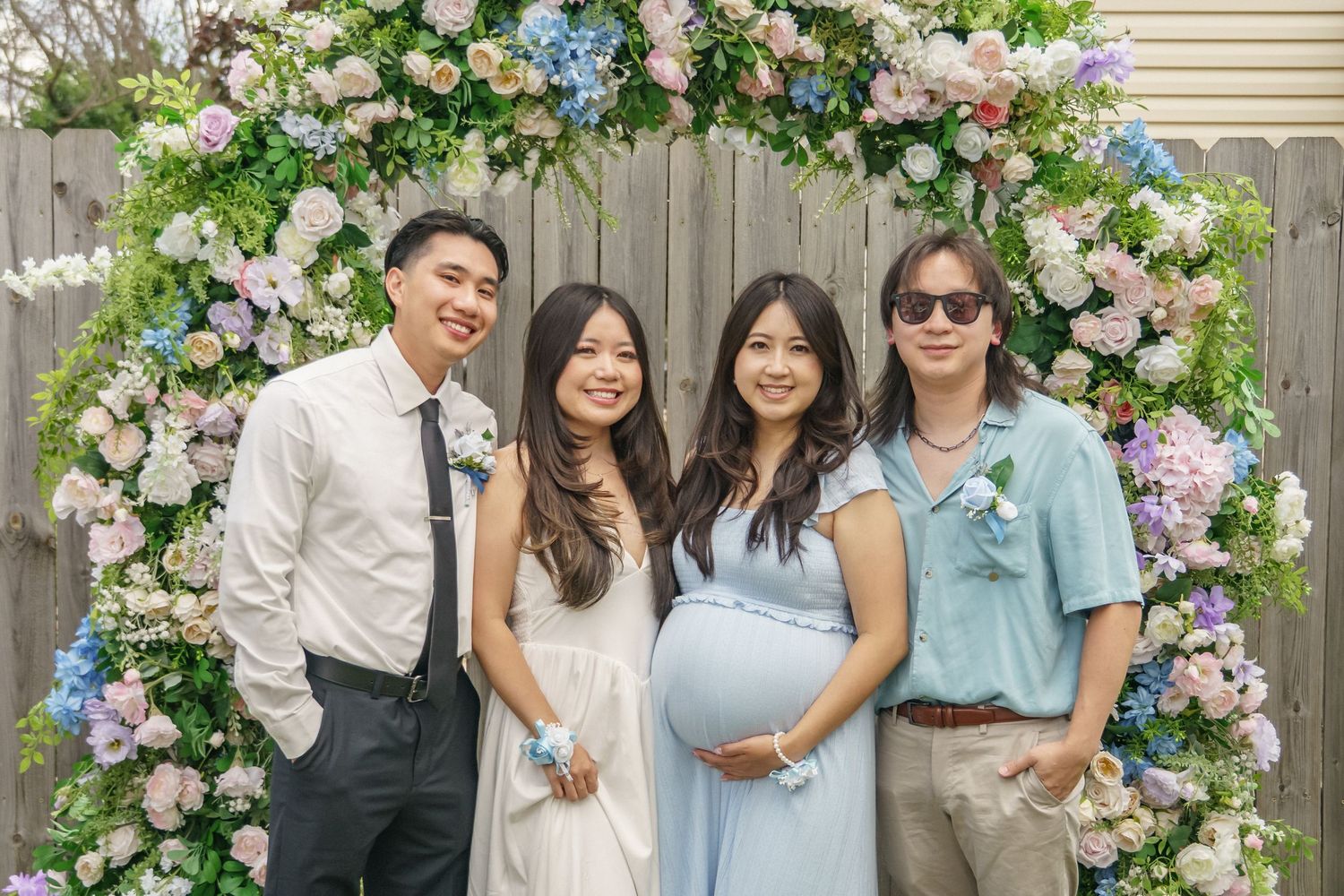 Four friends smile together under colorful floral arch at outdoor baby shower celebration.