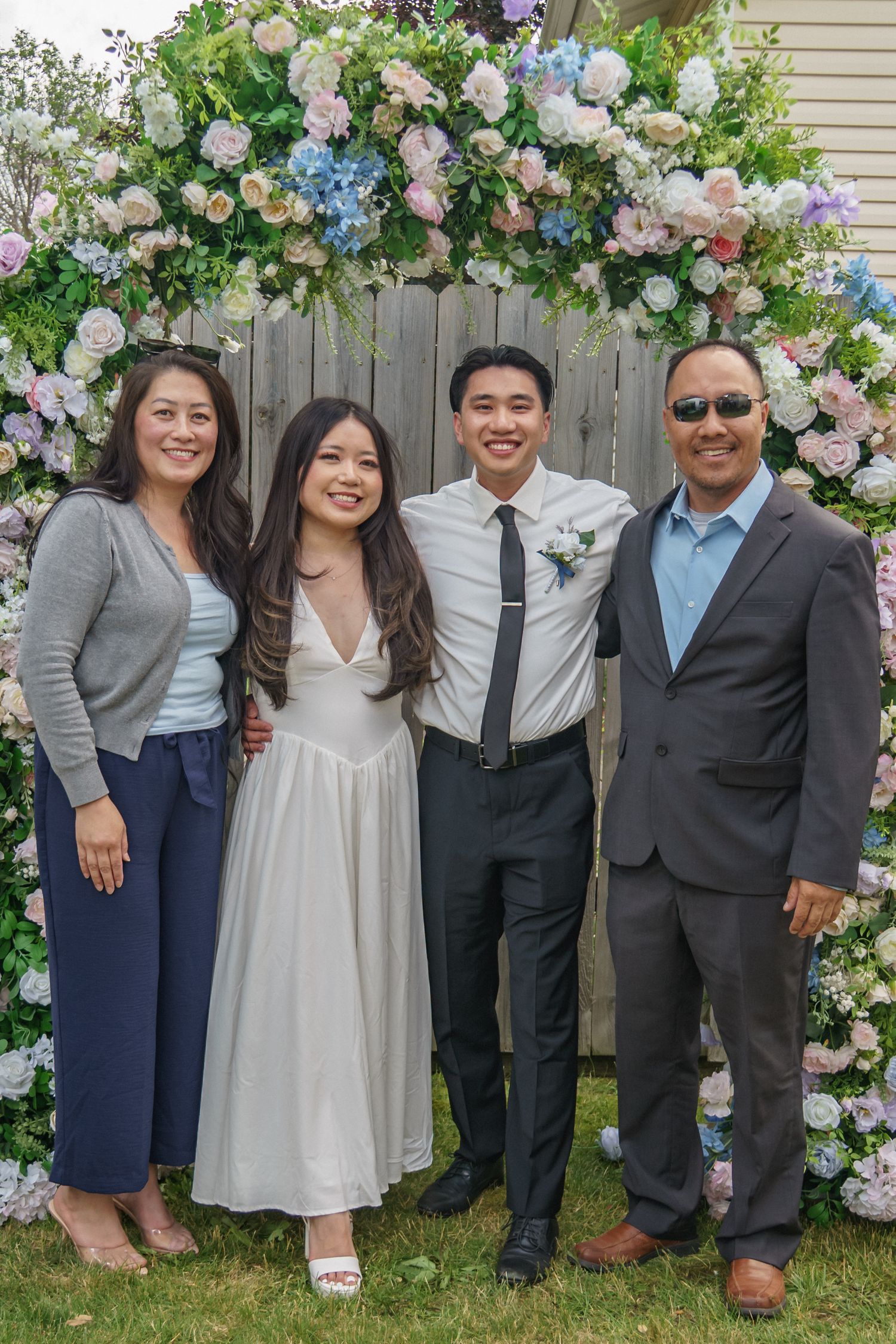 Family of four poses under a beautiful floral arch with pink and blue flowers at an outdoor wedding.
