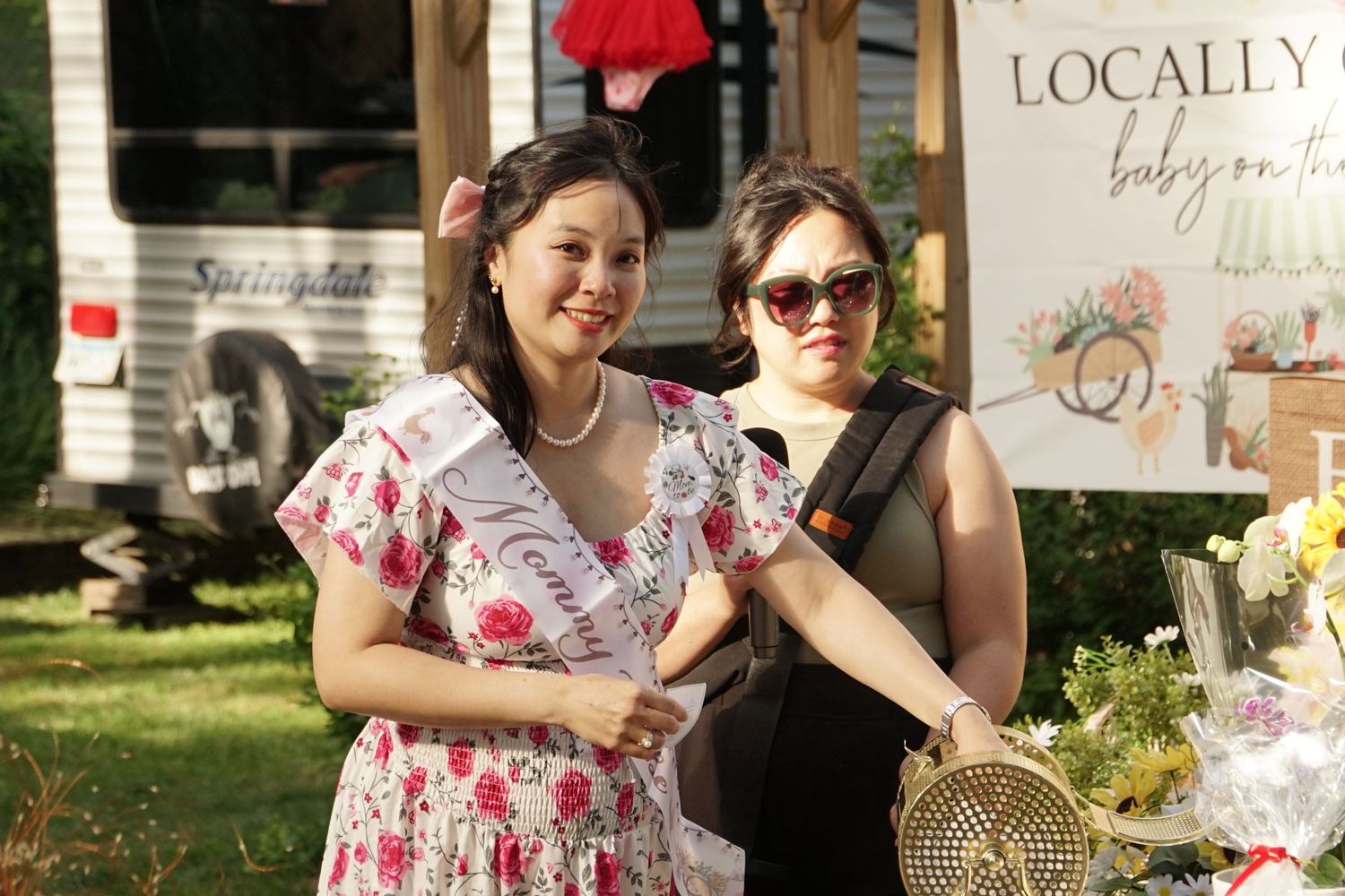 Two women pose outdoors at a local market, one wearing a floral dress with a pageant sash near a vendor booth.