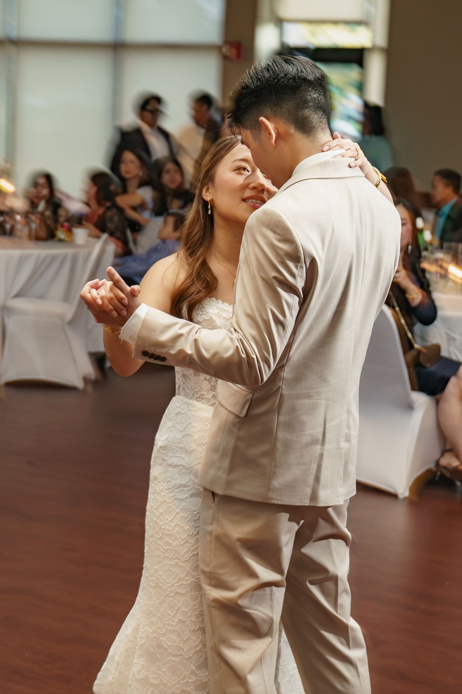 Bride and groom share their first dance at wedding reception, surrounded by seated guests at white-clothed tables.