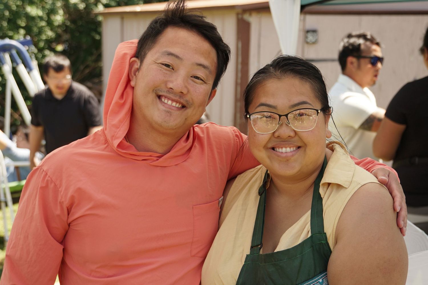 Two smiling friends pose together outdoors at a casual gathering on a sunny day.