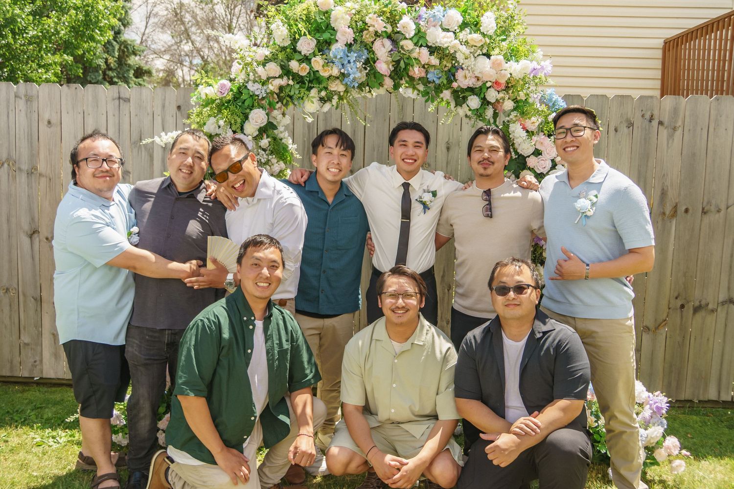 Group of ten men pose together in front of a flower arch at an outdoor backyard wedding ceremony.