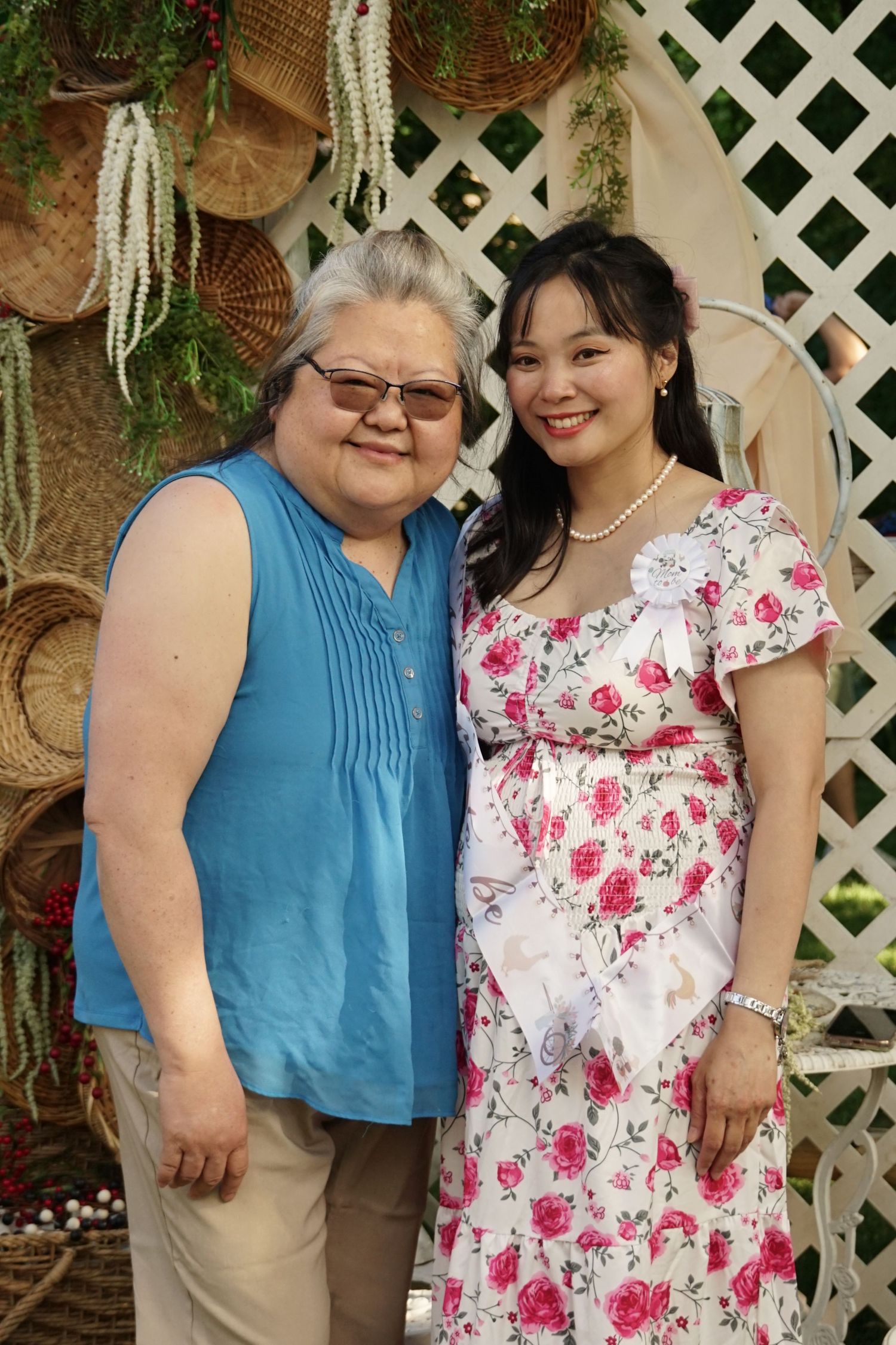 Two women smiling together outdoors, one in blue sleeveless top, other in floral dress, posed before white lattice backdrop.