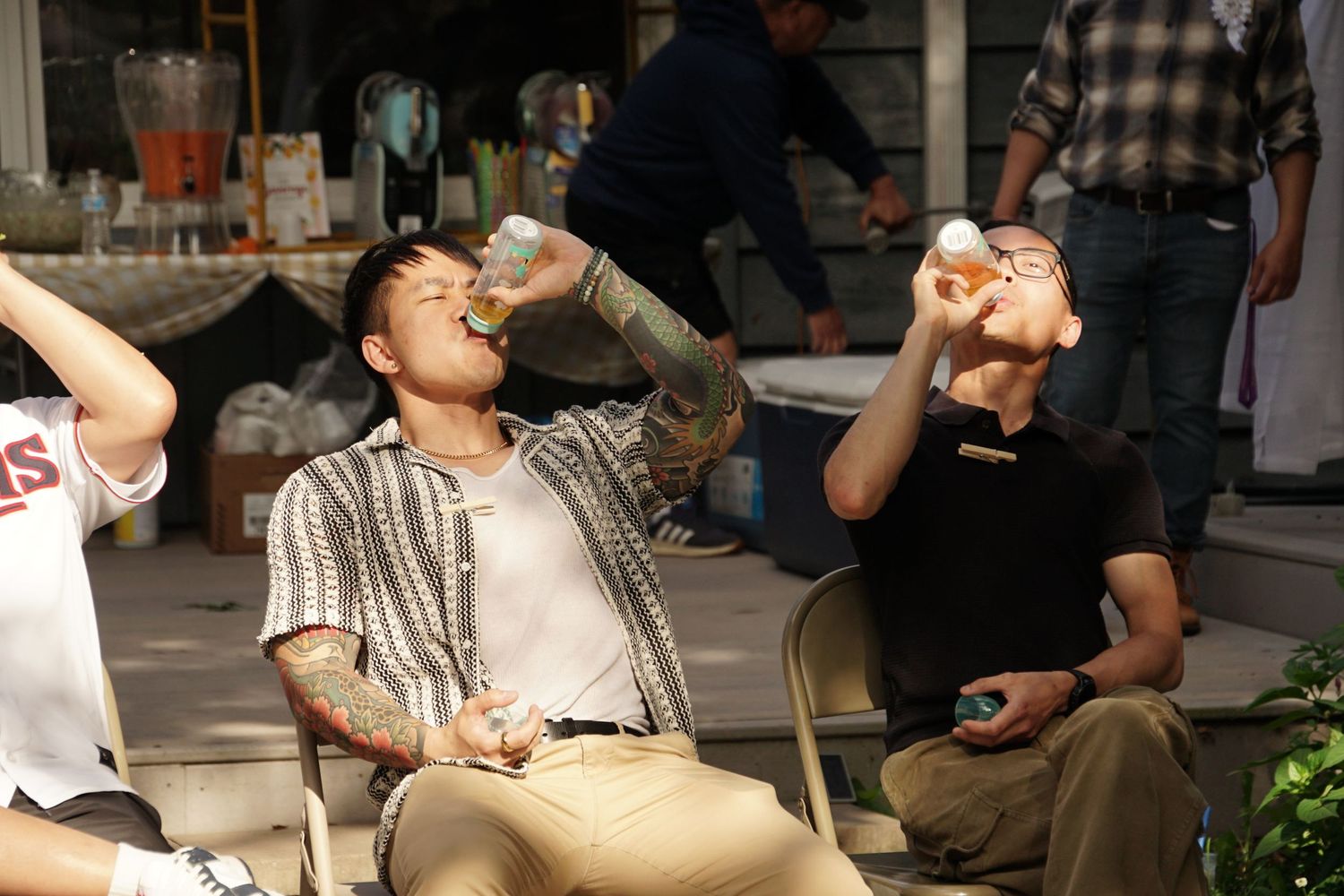 Two men sitting outdoors tipping back drinks at a casual gathering near a bar setup.