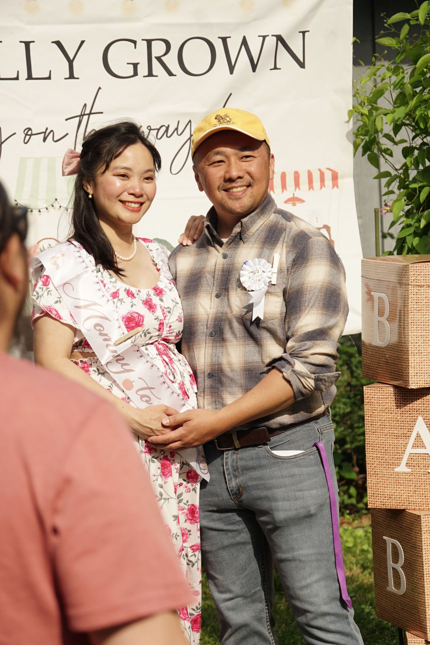 Pregnant couple smiling at baby shower, standing in front of a decorative banner outdoors.