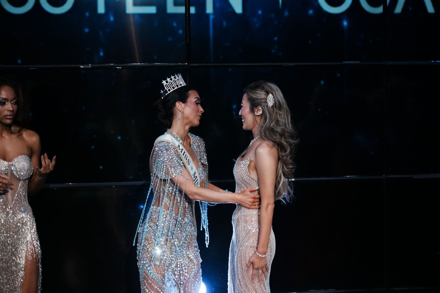 Two women in sparkling gowns share a moment as one wearing a crown places hands on the other at a pageant.