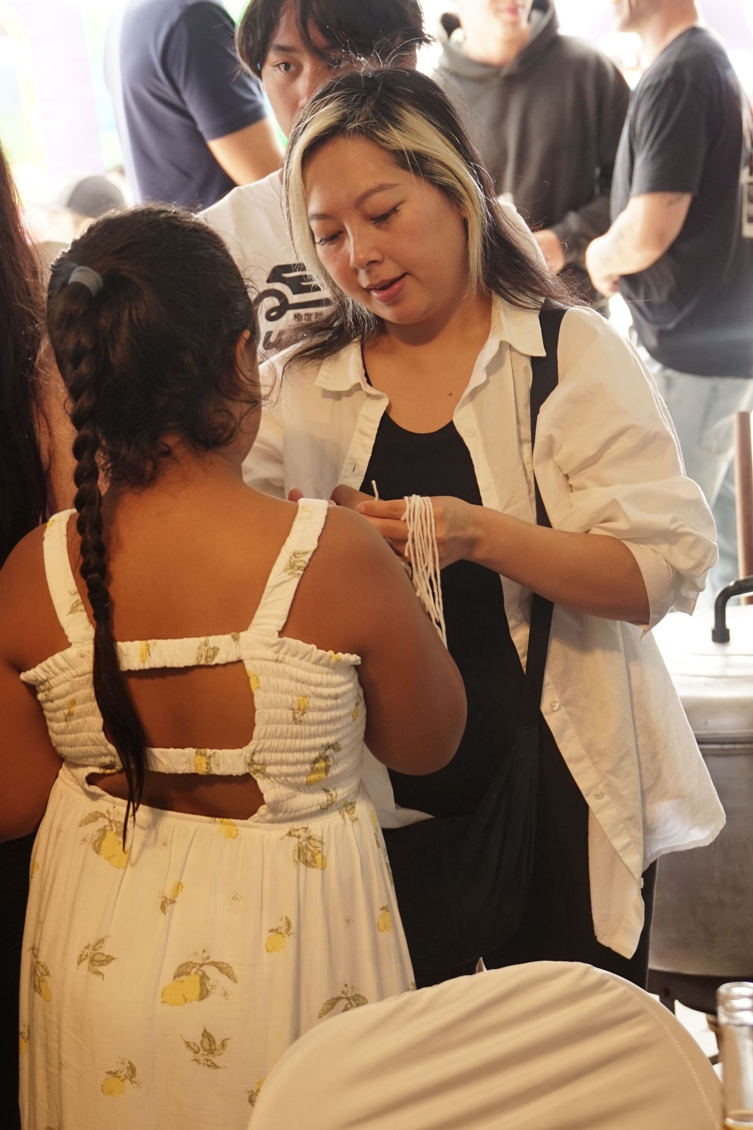 Woman in white shirt adjusting necklace on girl in floral dress with braided hair at indoor event.