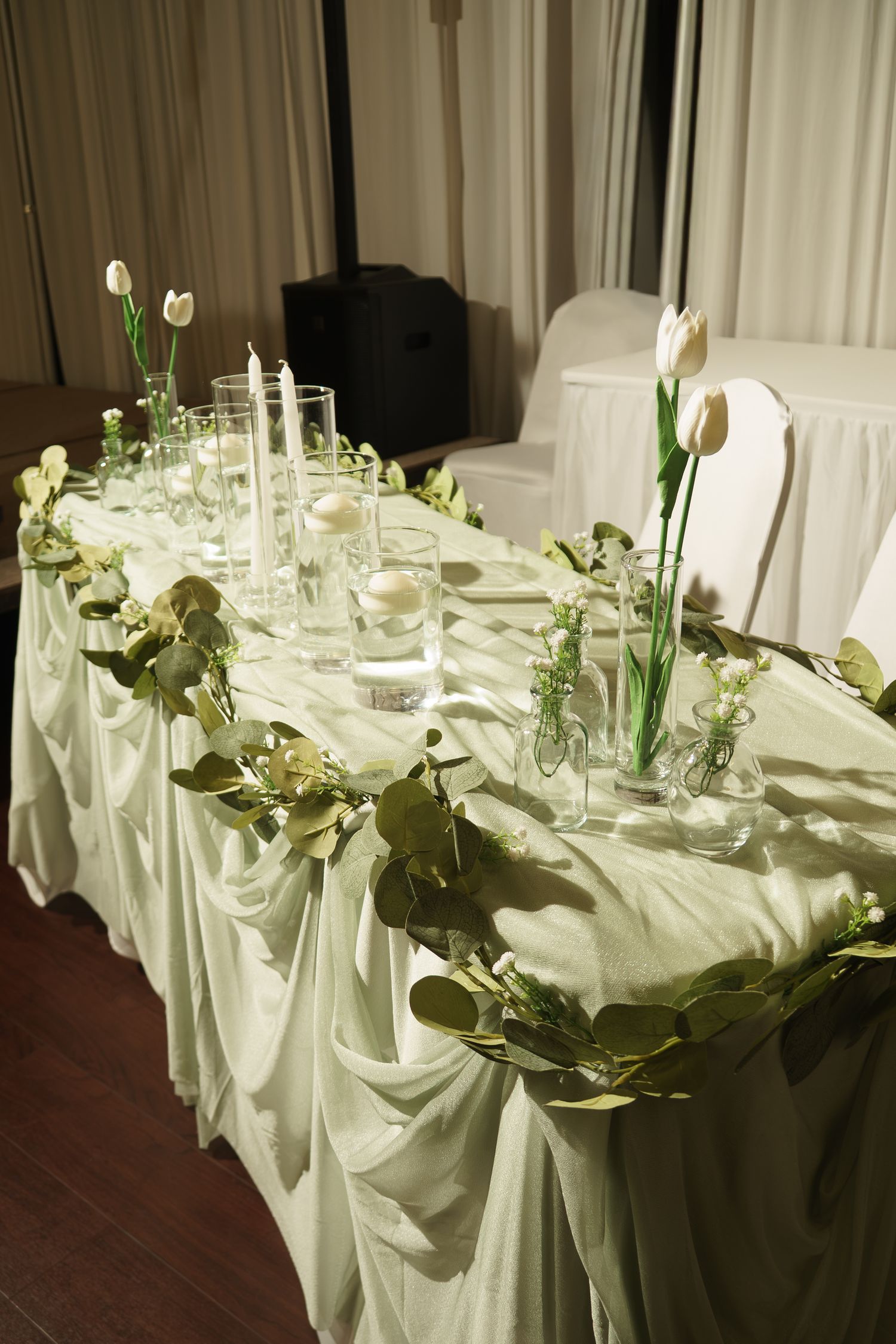 Elegant wedding head table with white tulips, greenery garland, candles, and draped white fabric skirting.