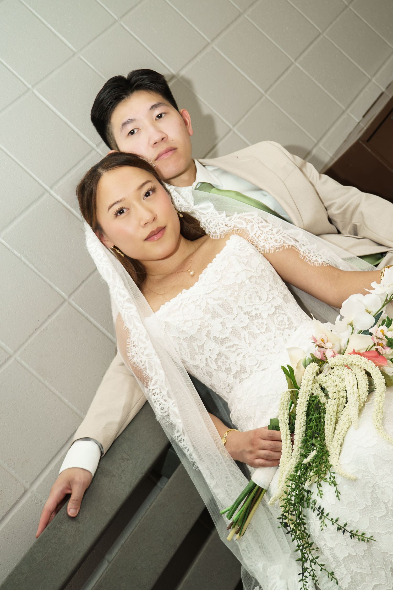 Asian bride and groom in wedding attire pose together, bride holding floral bouquet with white veil, groom in beige suit.