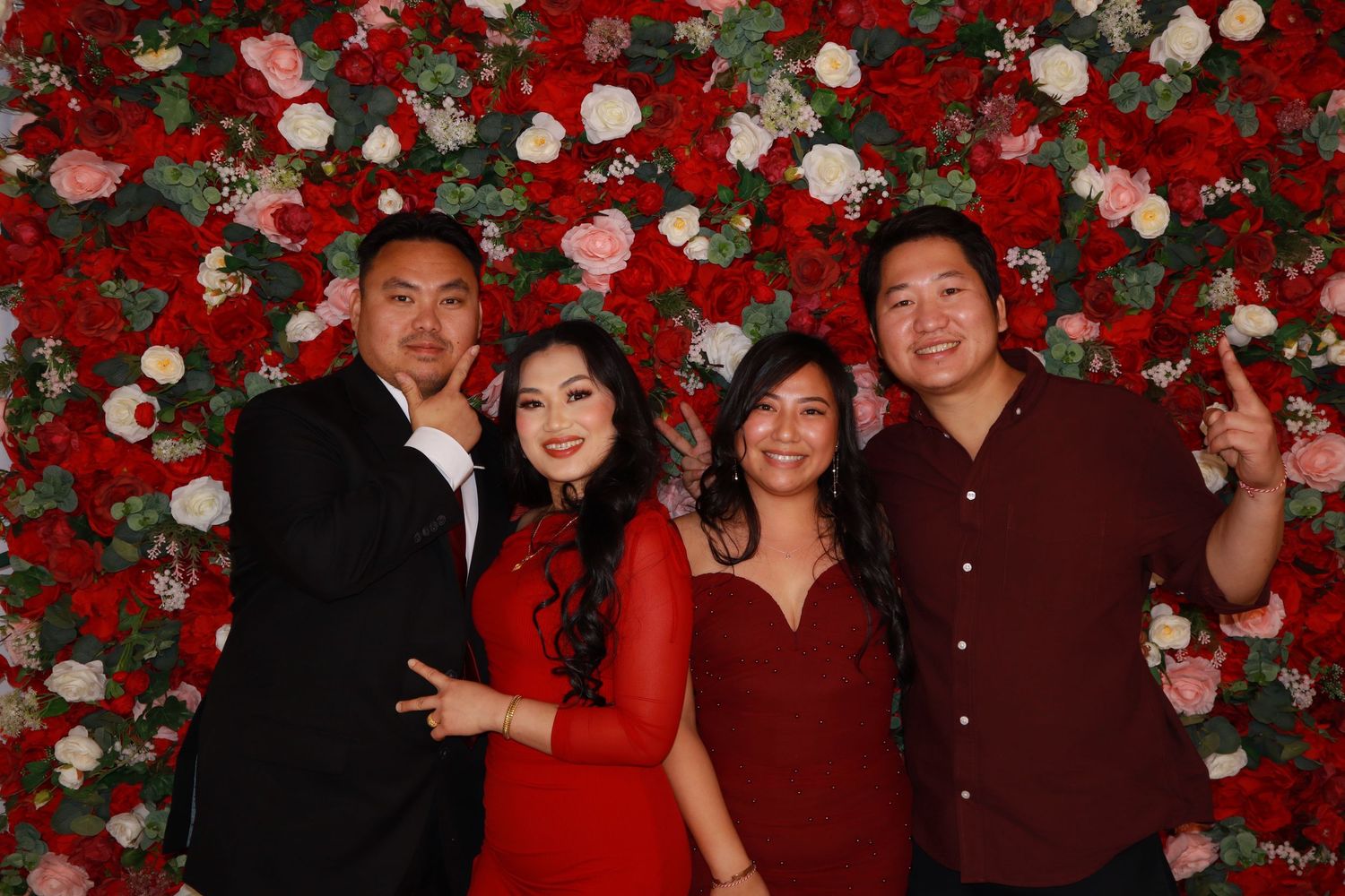 Four people in red and black outfits smiling in front of a floral rose wall backdrop.