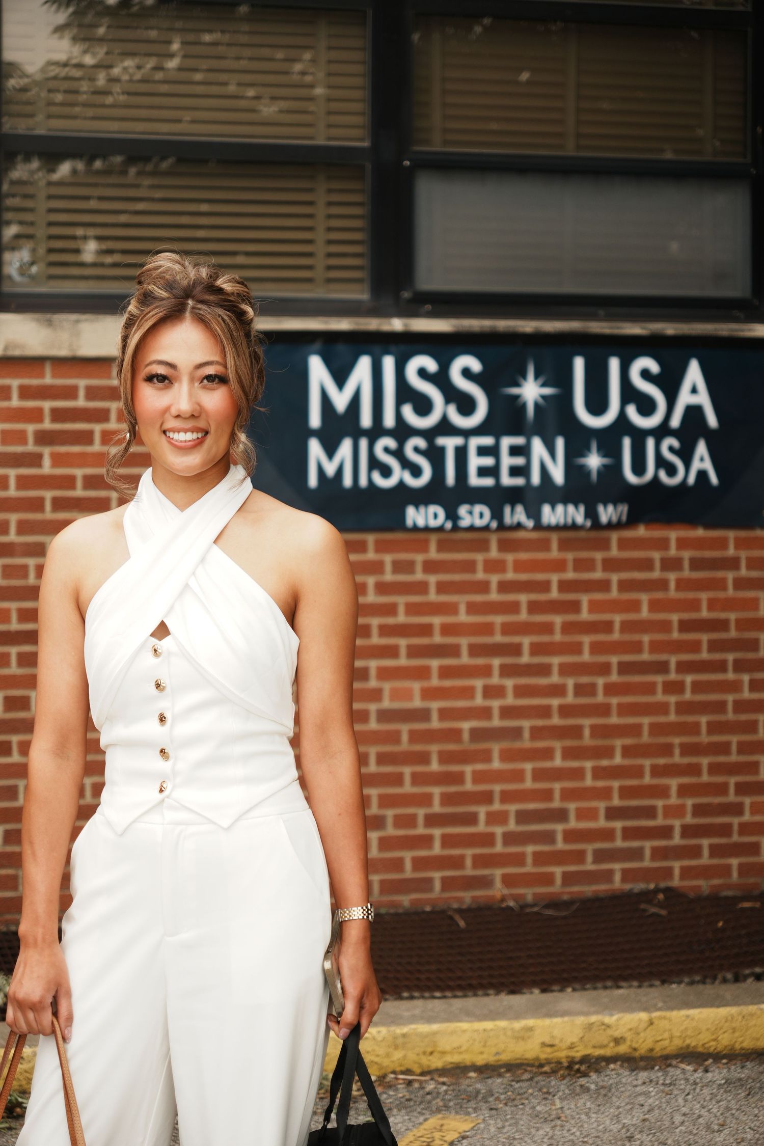 Smiling woman in white halter jumpsuit standing in front of Miss USA and Miss Teen USA brick building sign.