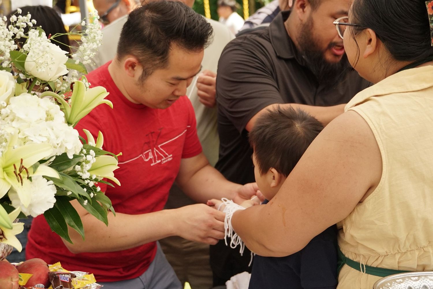 Adults gather around a baby during a cultural ceremony with white floral arrangements on a decorated table.