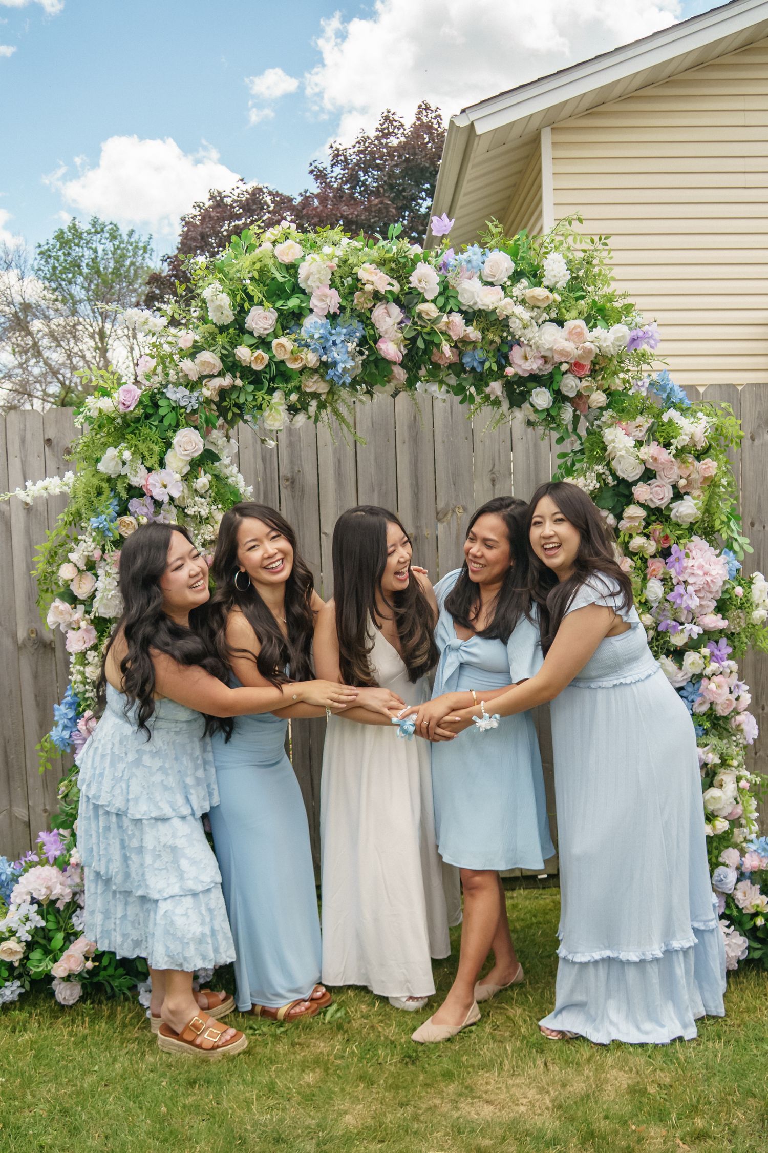 Five bridesmaids in blue dresses pose under a floral arch with pink and white blooms at an outdoor wedding.