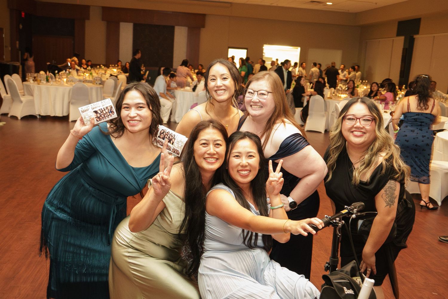Six women posing together at a formal banquet event, smiling and holding small signs in an elegant ballroom.