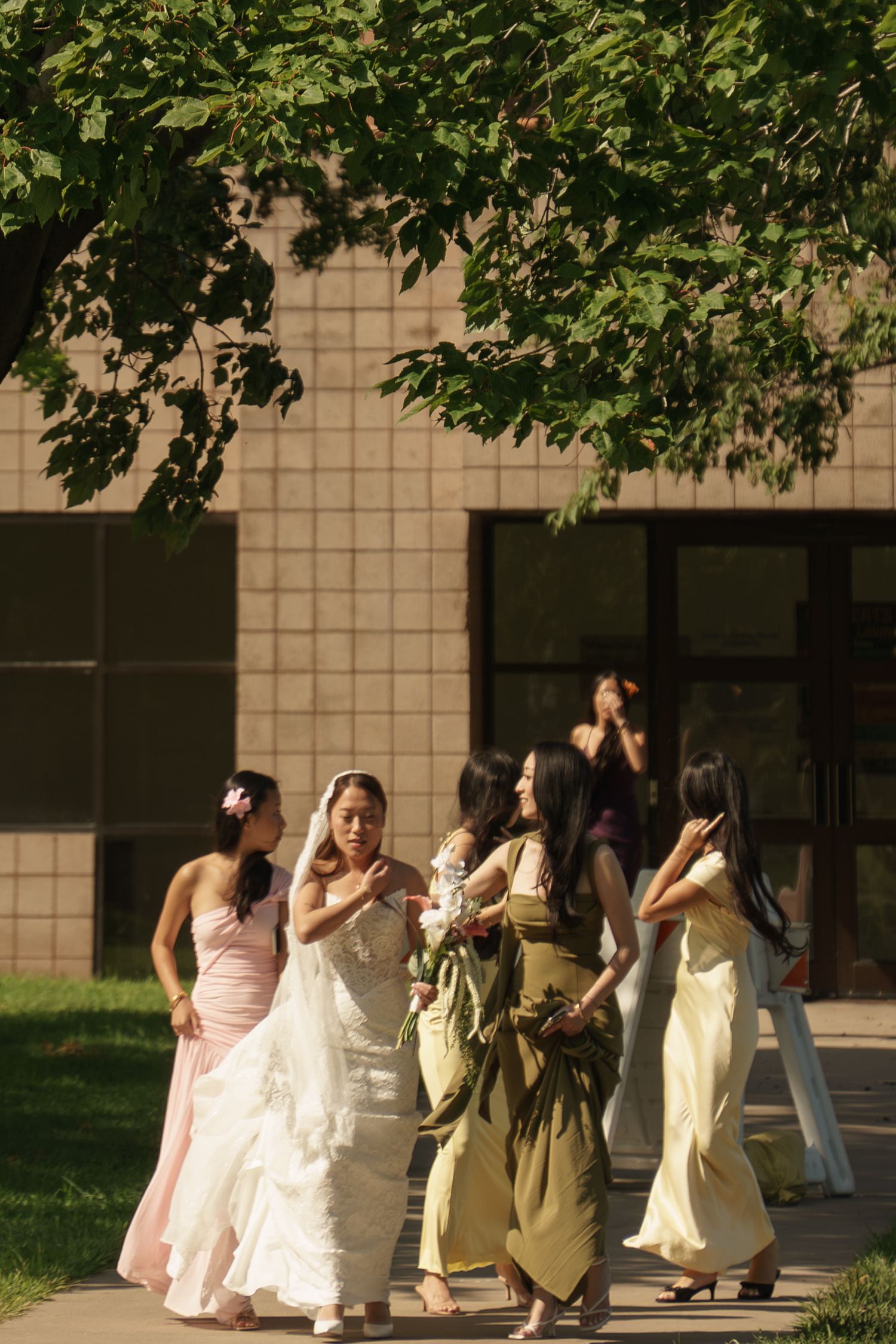 A bride in white gown walks with bridesmaids in pastel dresses outdoors near a building on a sunny day.