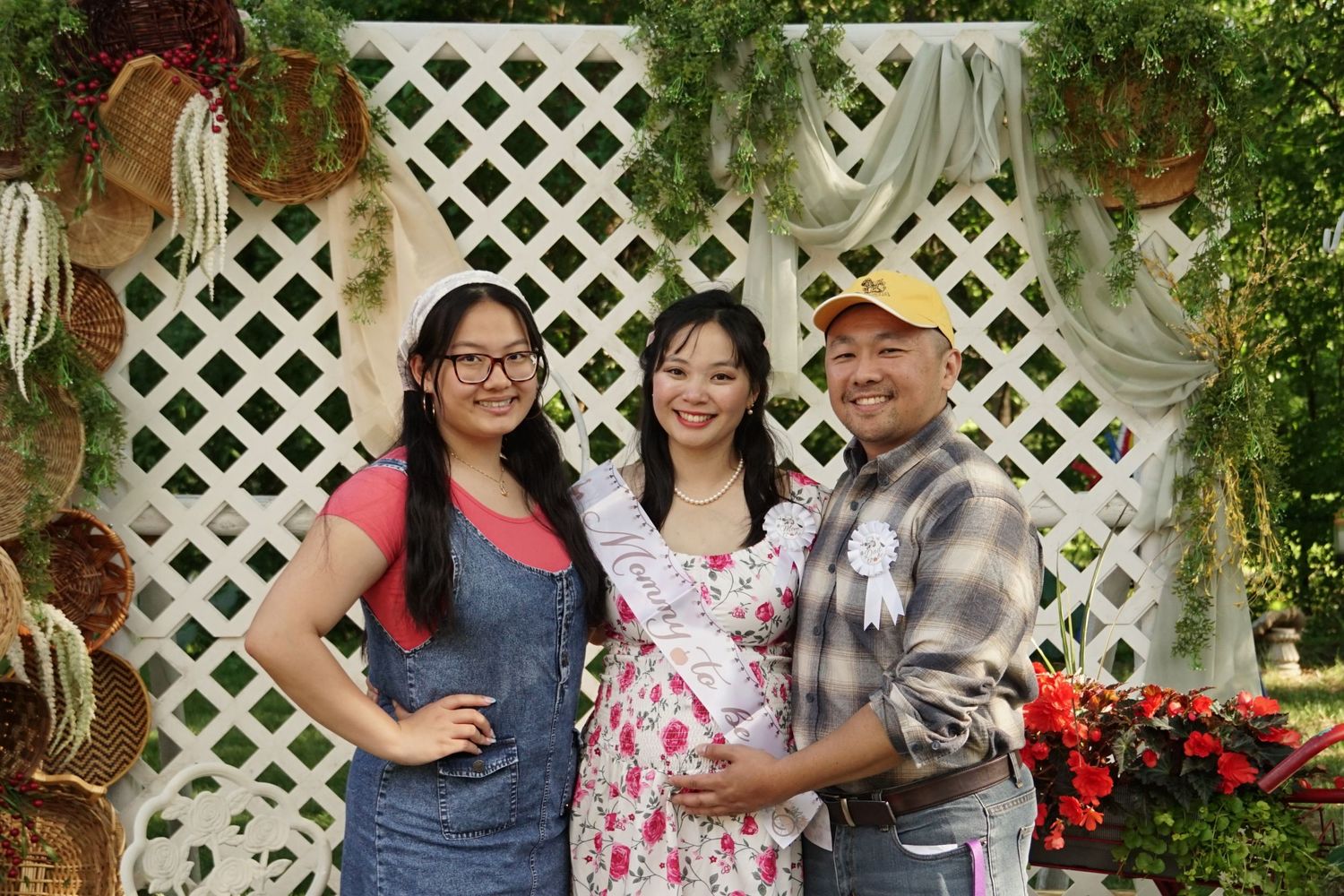 Smiling Asian family of three posing in front of white lattice backdrop with greenery, pregnant woman in floral dress.