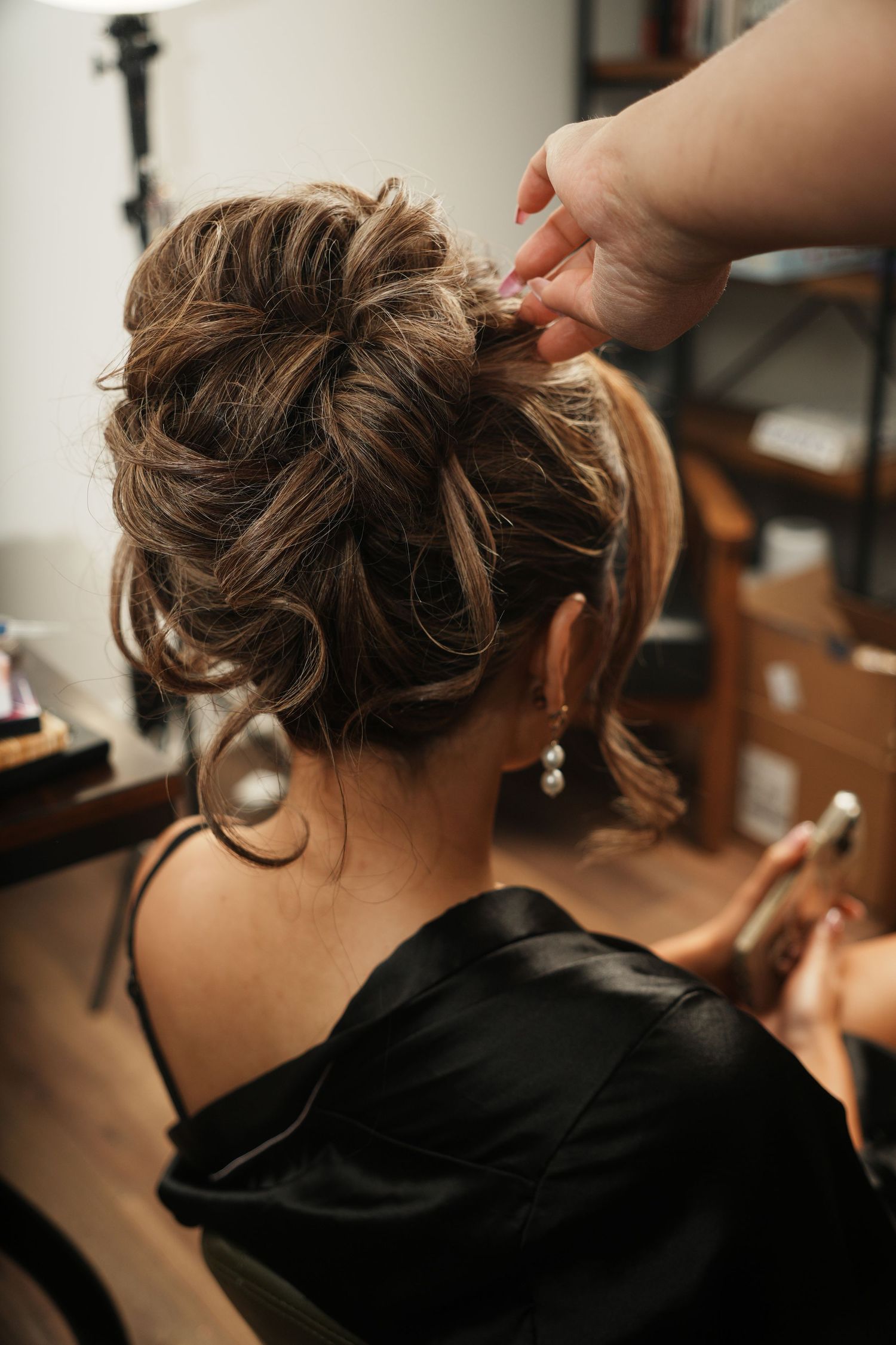 Hairstylist pinning an elegant updo on a woman wearing a black satin robe, with pearl drop earrings visible.