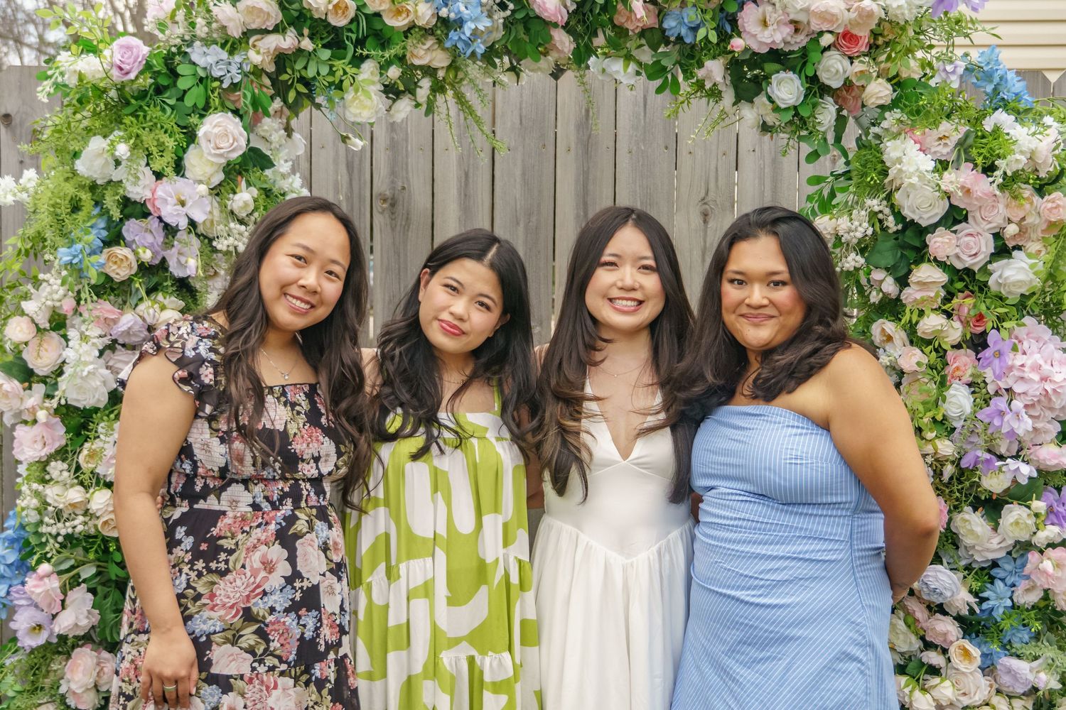 Four women smile together under a lush floral arch with pink and blue flowers at a wedding celebration.