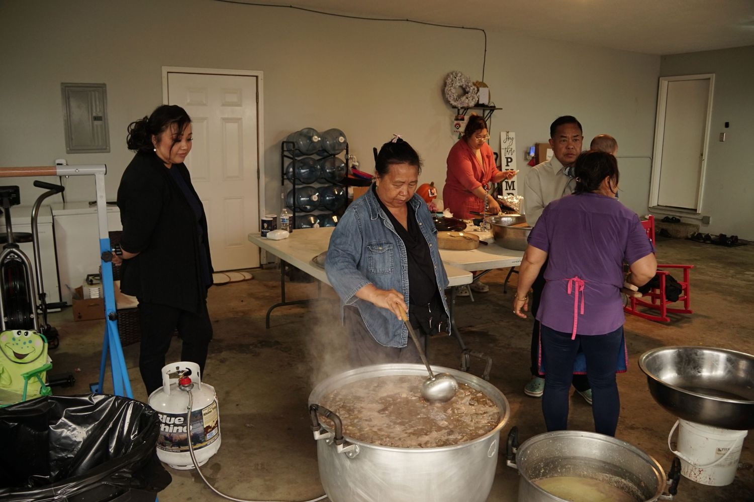 Women cooking large pots of food in a community kitchen, stirring steaming dishes during a group meal preparation event.