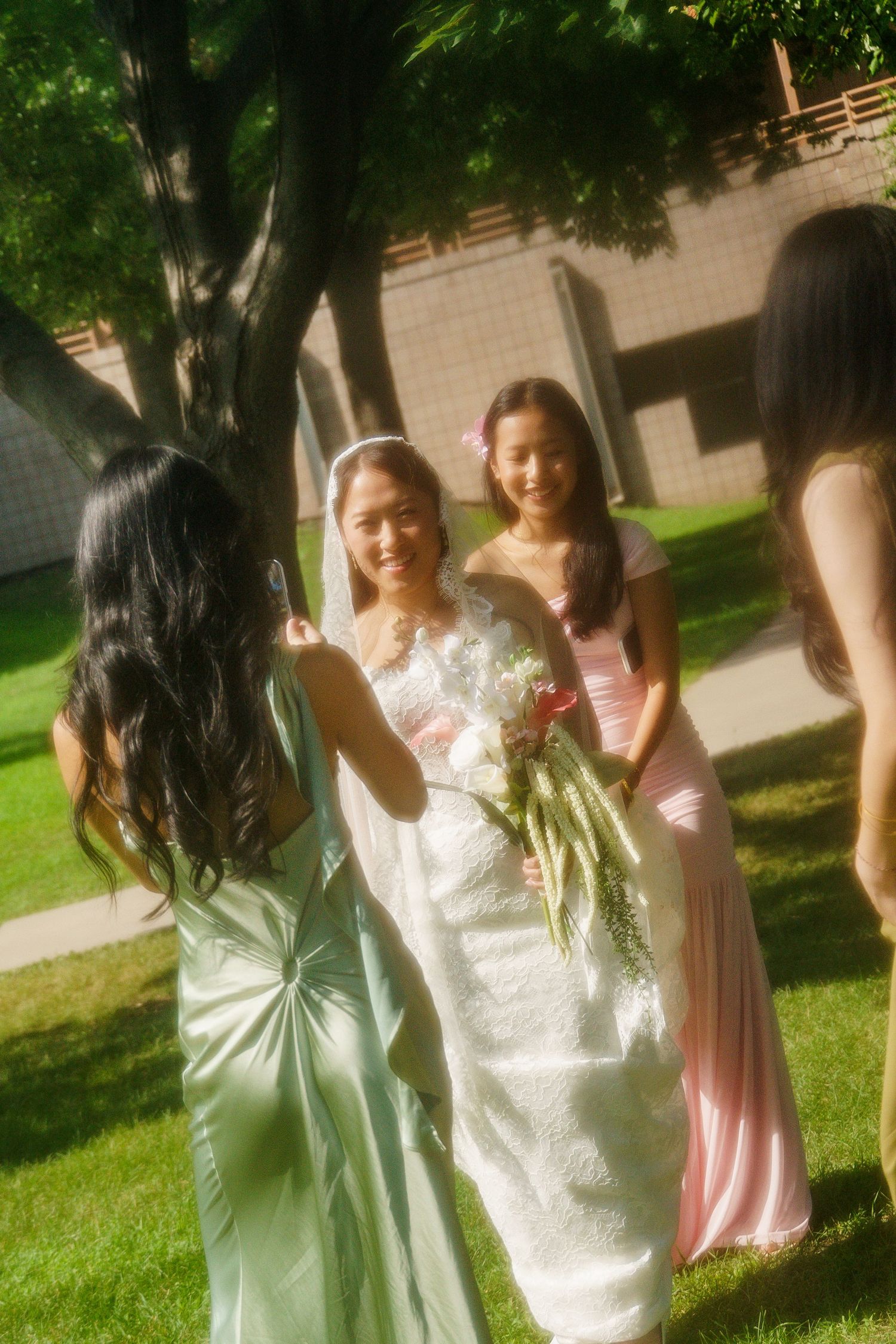 A smiling bride in white holds bouquet with bridesmaids in green and pink dresses outdoors on green lawn.