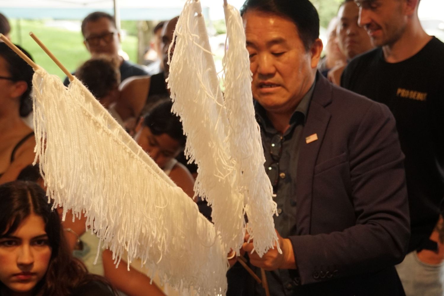A man in a blue blazer holds white fringed fabric at a crowded cultural event outdoors.