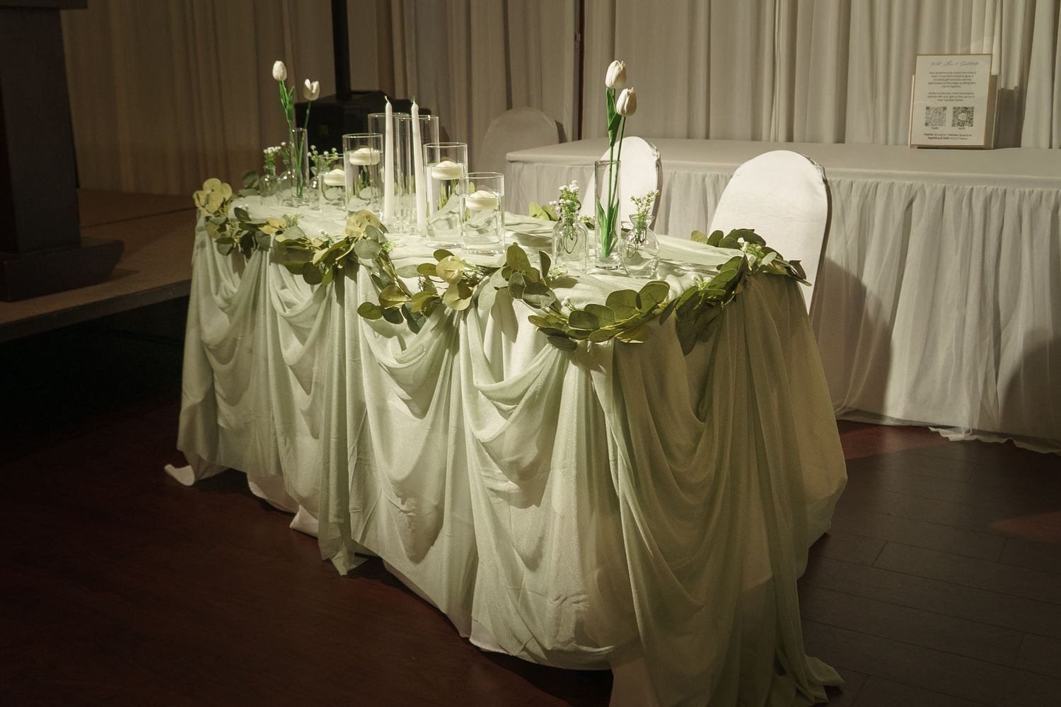 Elegant wedding head table with white draped fabric, green garland, candles, and white calla lily floral arrangements.